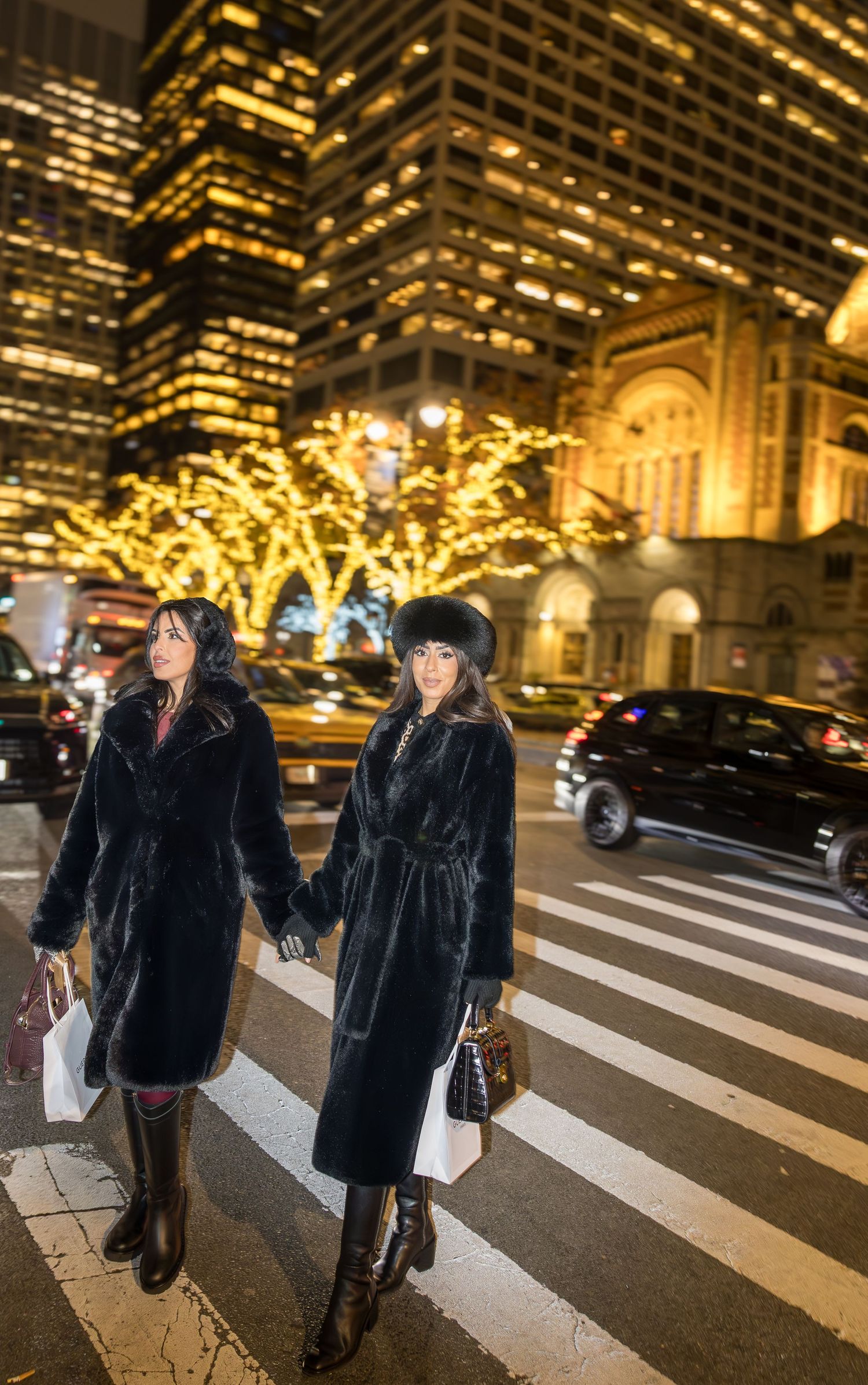 Two elegant women in matching black fur coats hold hands crossing a city street amid dazzling holiday lights.