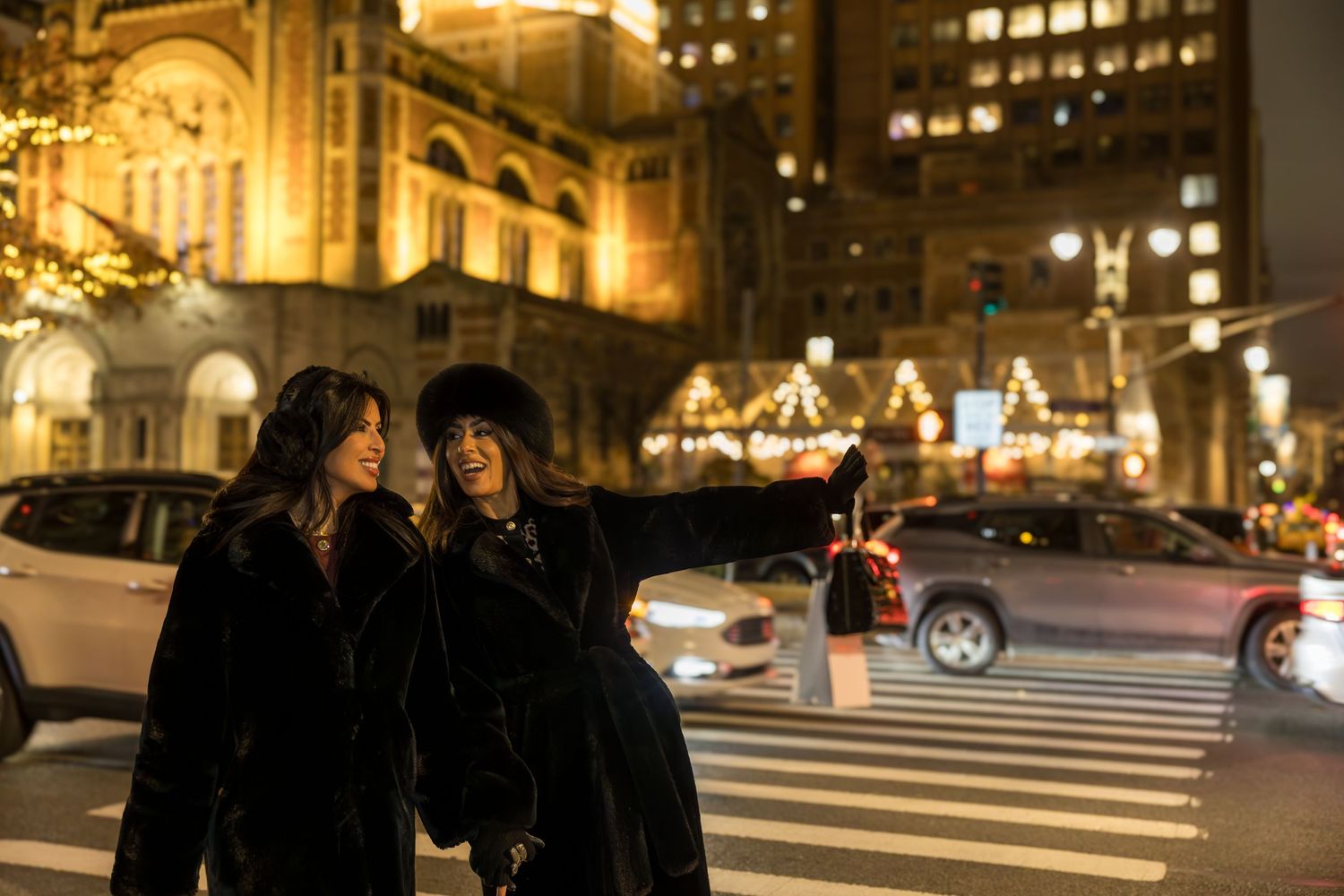 Two women in black fur coats stand on a festive night street, one pointing into the distance at city lights.