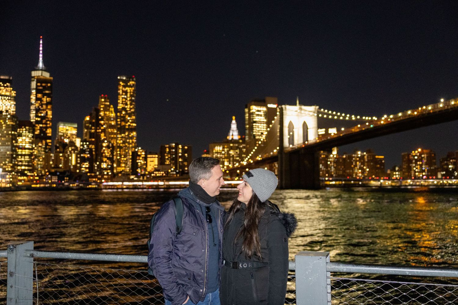 Couple kissing near Brooklyn Bridge with Manhattan skyline glowing at night.