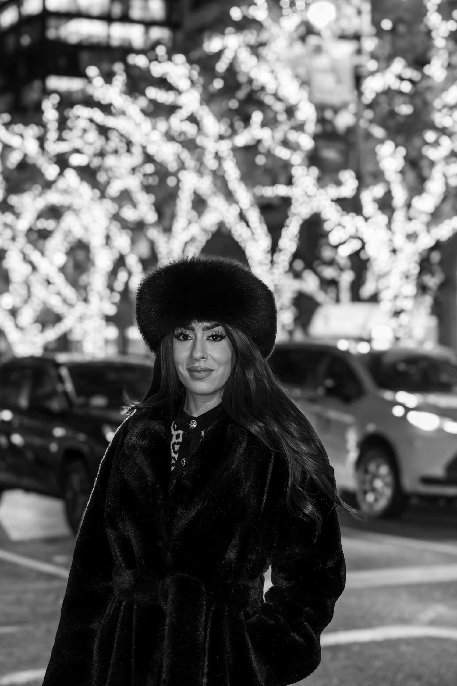 Black and white portrait of a woman in fur coat and hat posing before bokeh holiday lights on a city street.