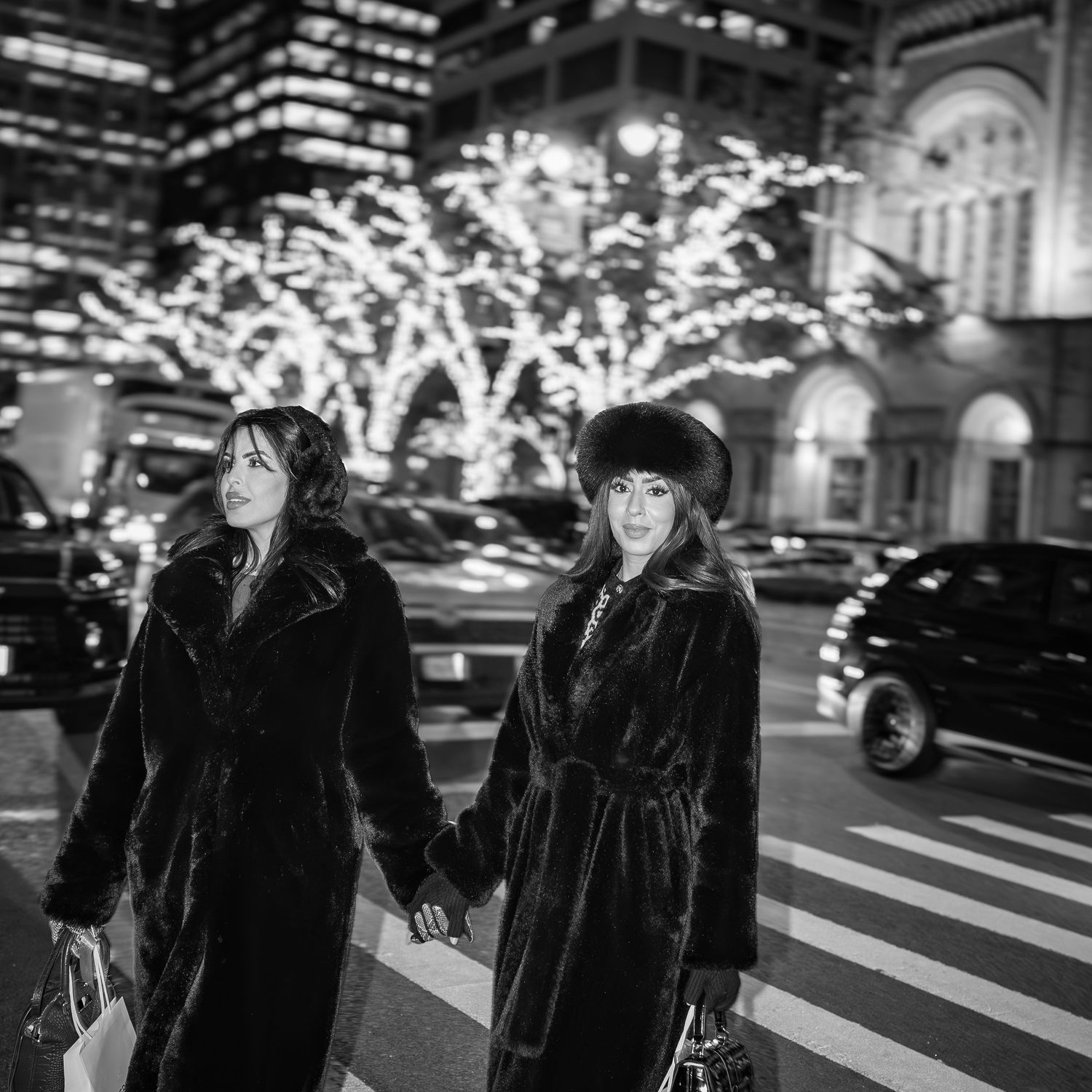 Black and white photo of two women in black fur coats holding hands crossing a street with holiday lights.