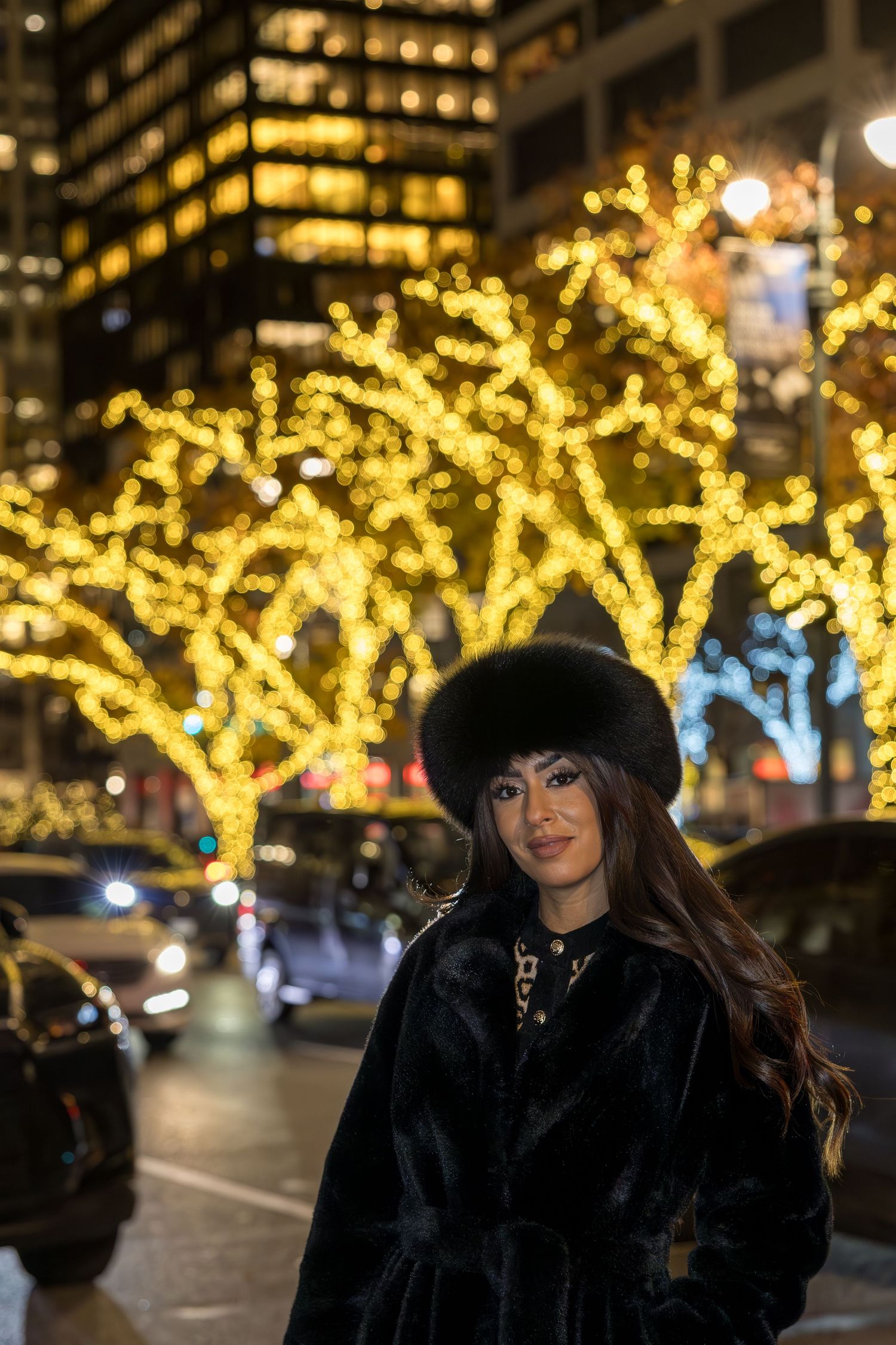 Elegant woman in black fur coat and fur hat poses before golden illuminated trees on a city street at night.