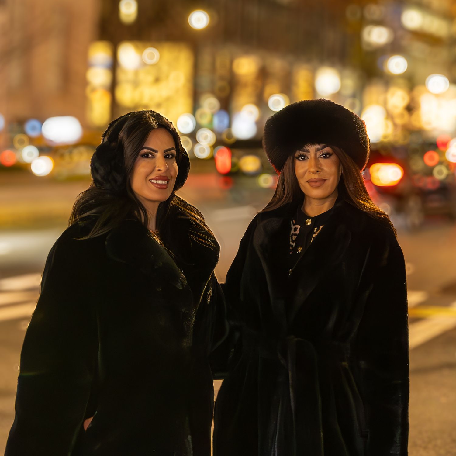 Two women in matching black fur coats posing on a city street at night with bokeh lights behind them.