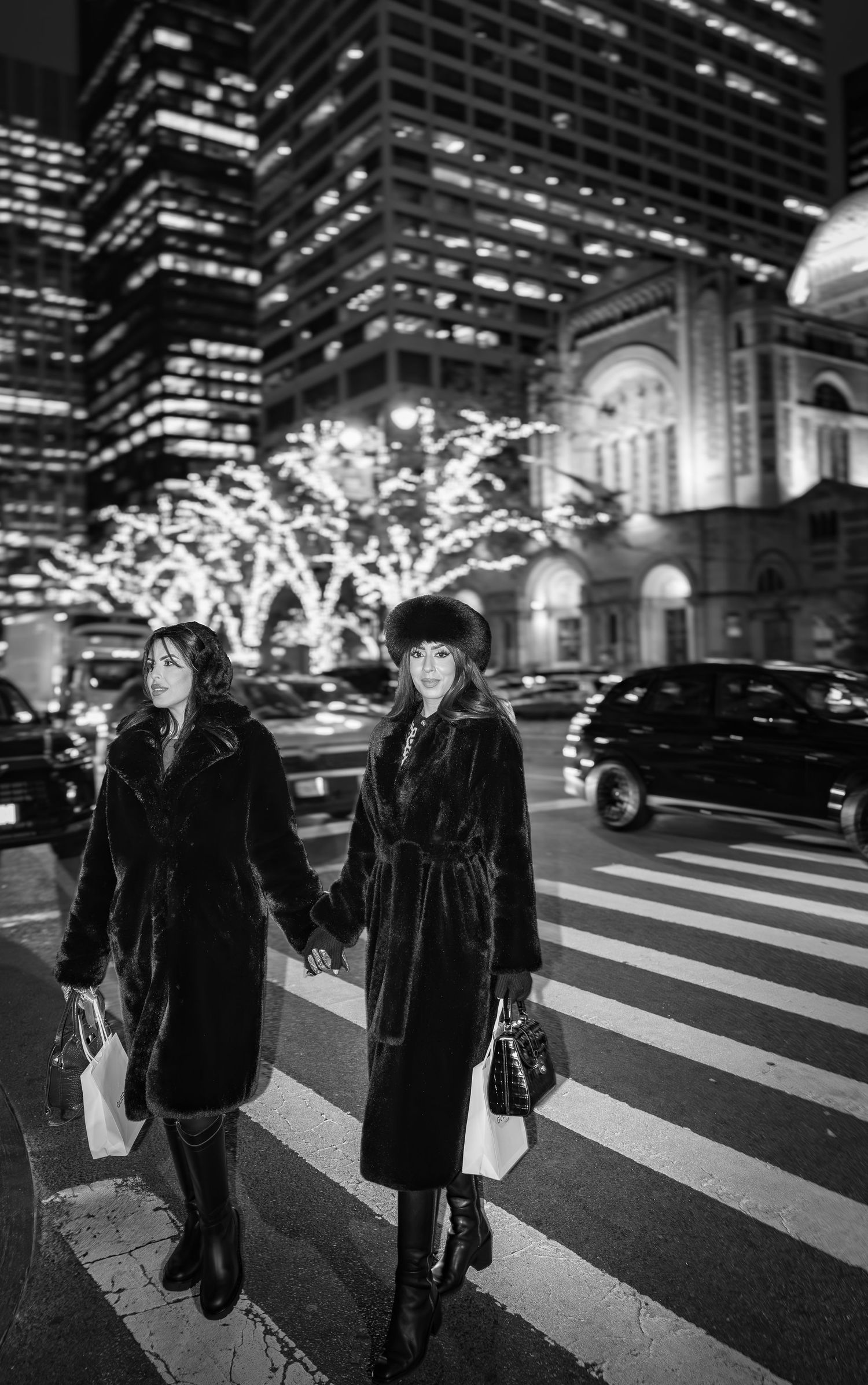 Two elegantly dressed women in long black coats holding hands while crossing a city street at night.