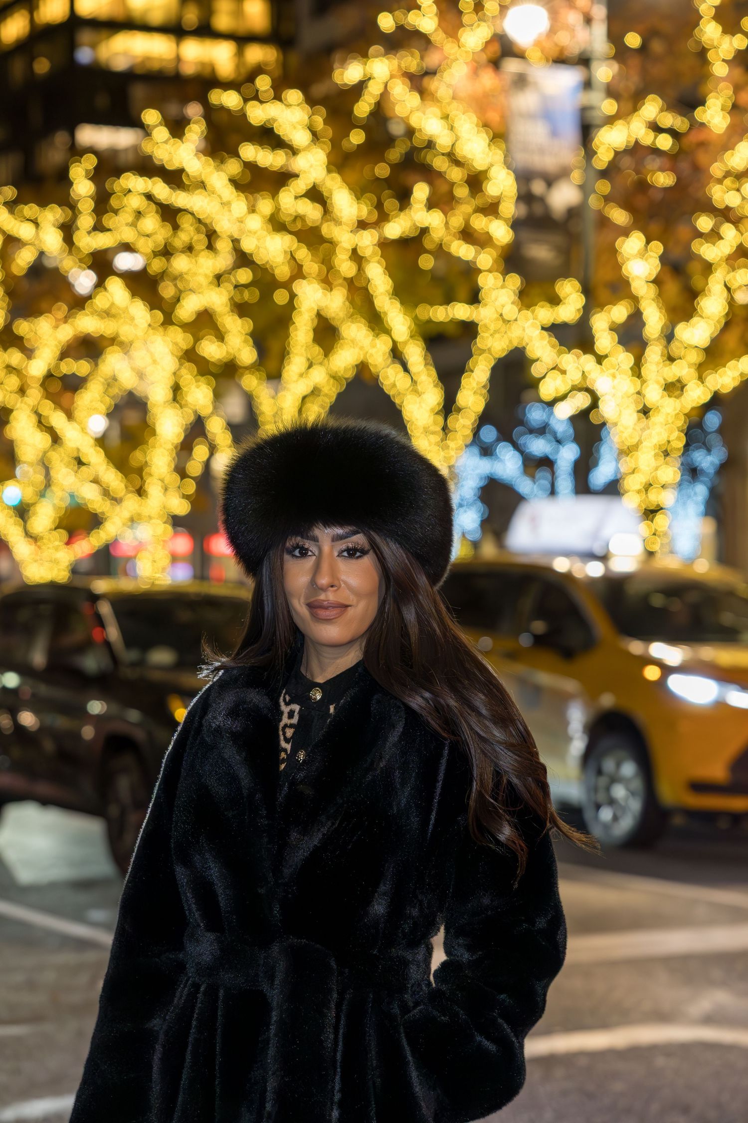 Stylish woman in black fur coat and oversized fur hat stands before sparkling golden holiday lights at night.
