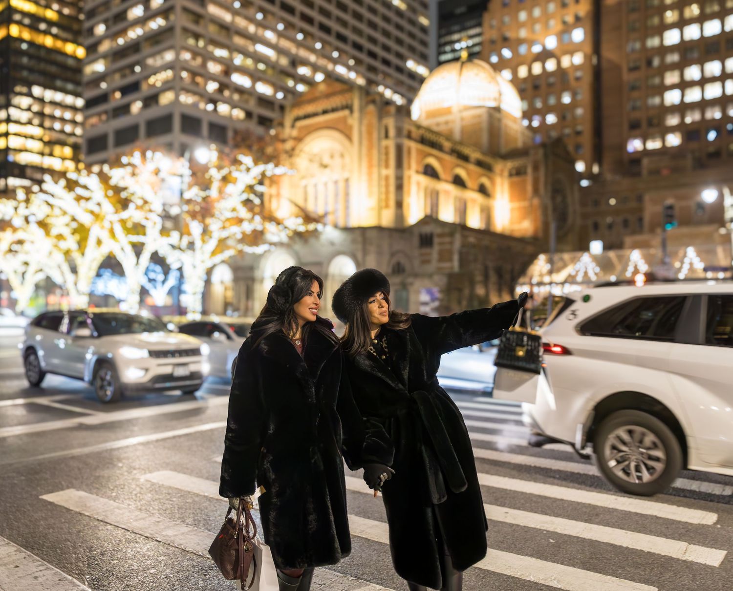 Two women in black fur coats stand on a festive city street at night, one pointing toward bright city lights.