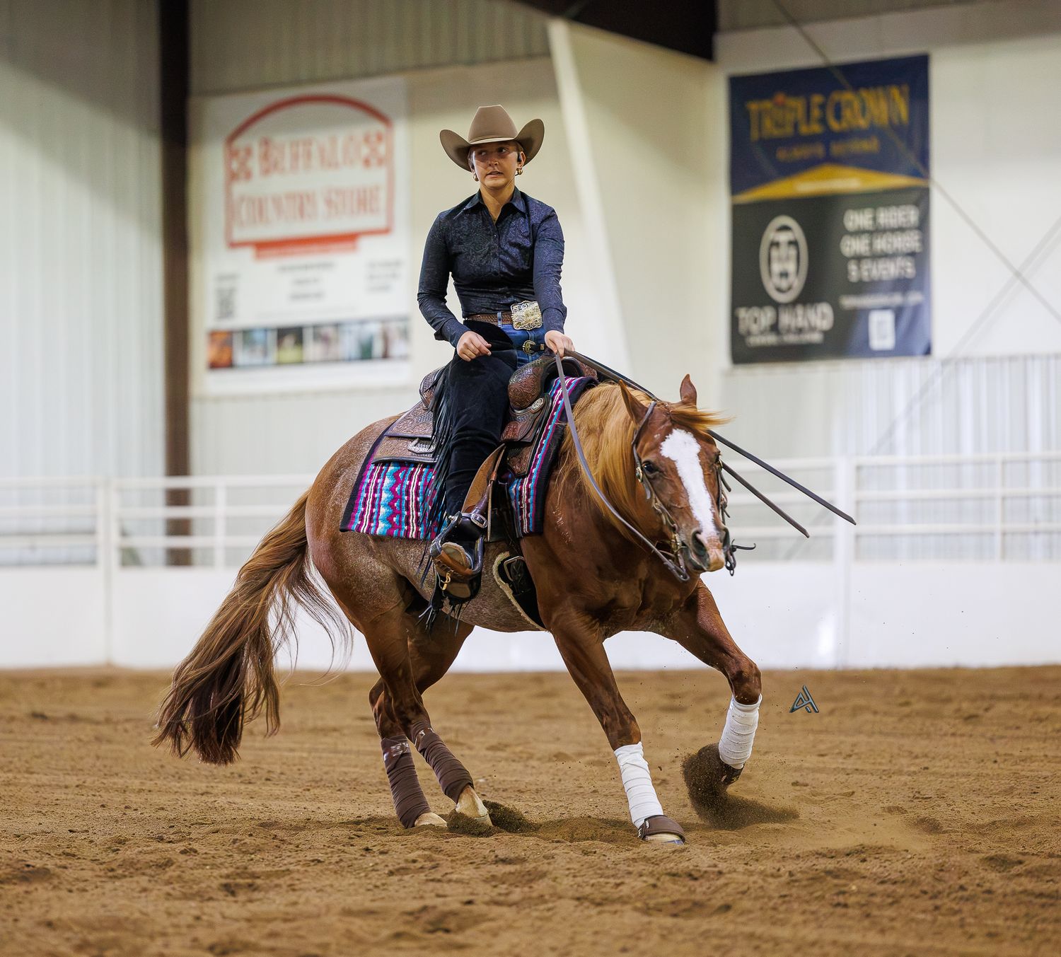 A female rider in a cowboy hat competes on a chestnut horse in a western reining event indoors.