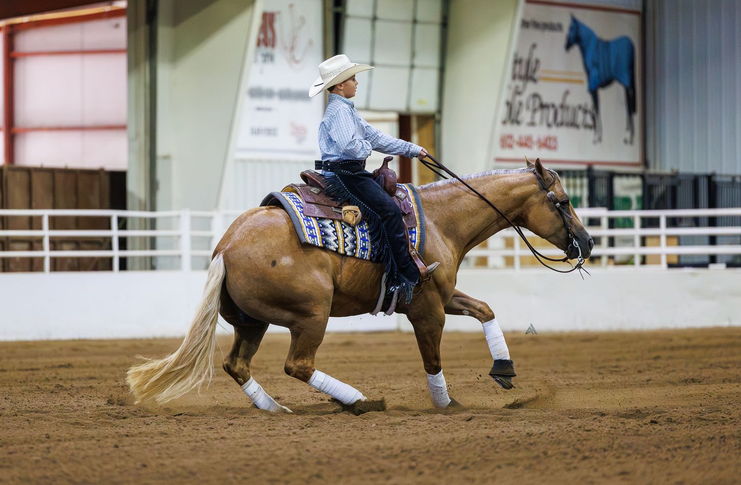 A rider in western attire on a palomino horse competing in an indoor equestrian arena on a dirt floor.