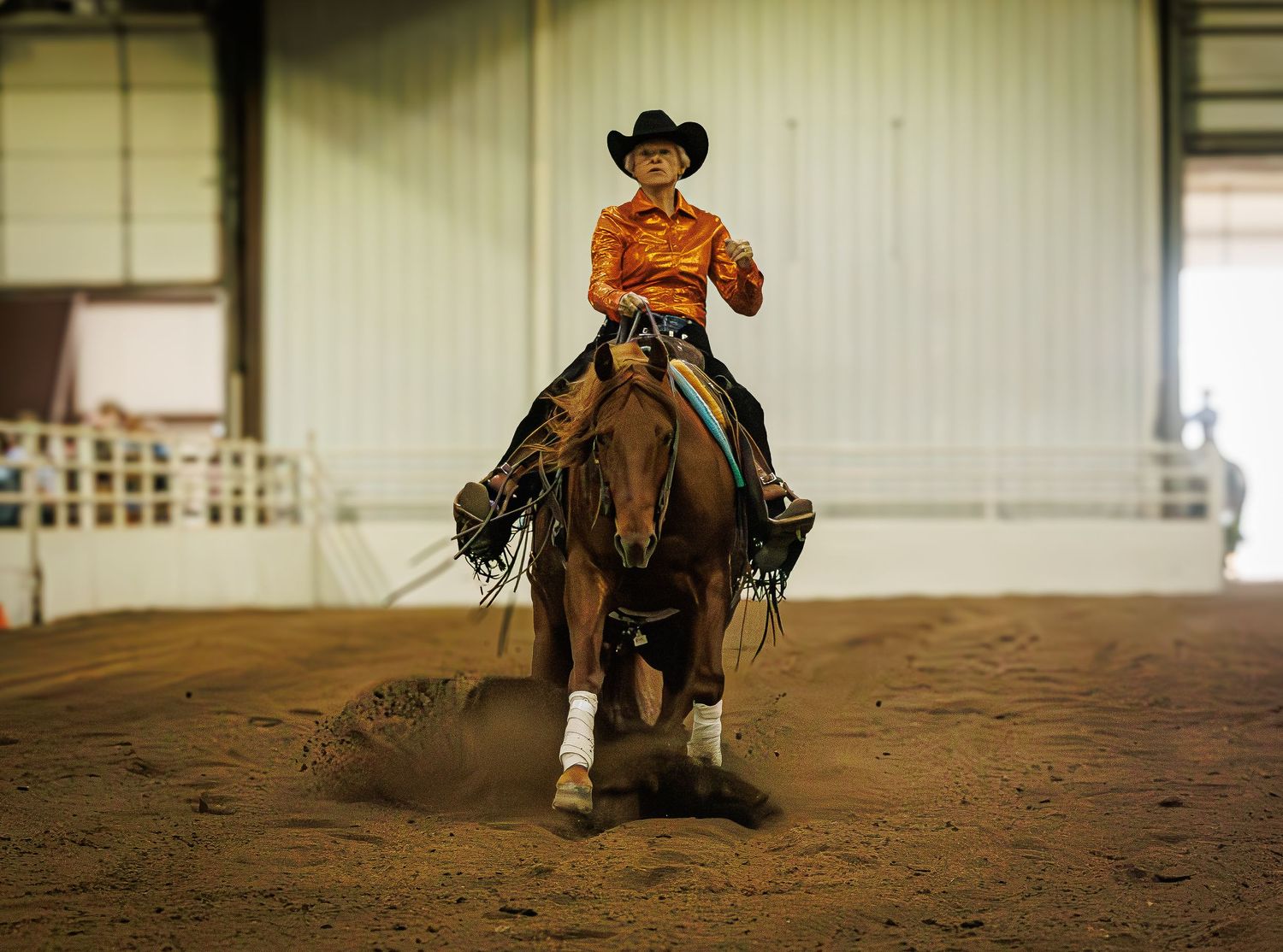 A cowboy in an orange shirt and black hat performs a reining maneuver on a chestnut horse in an indoor arena.