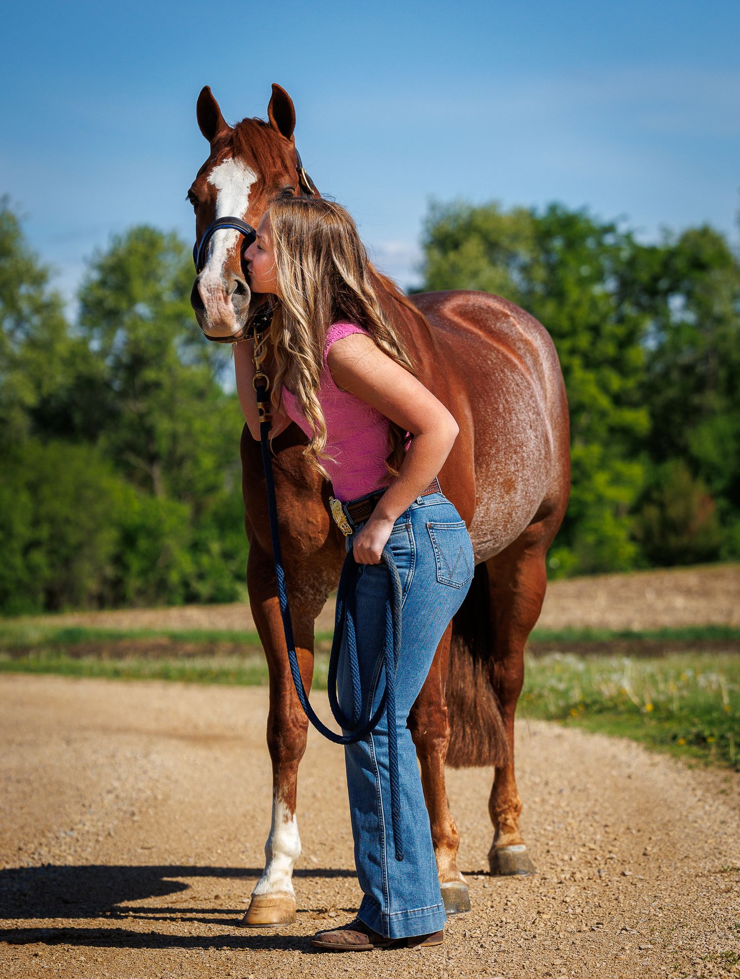 Young woman in jeans and pink top kissing a chestnut horse on a dirt path with green trees behind.