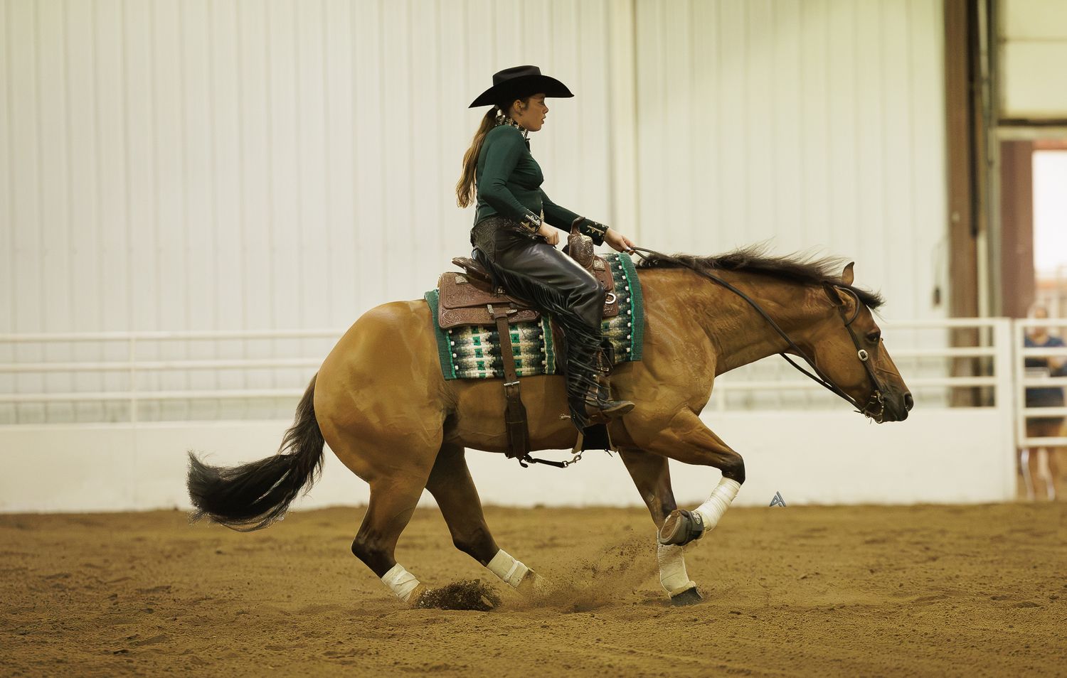 Woman in black cowboy hat riding a golden horse in an indoor equestrian arena during a western riding competition.