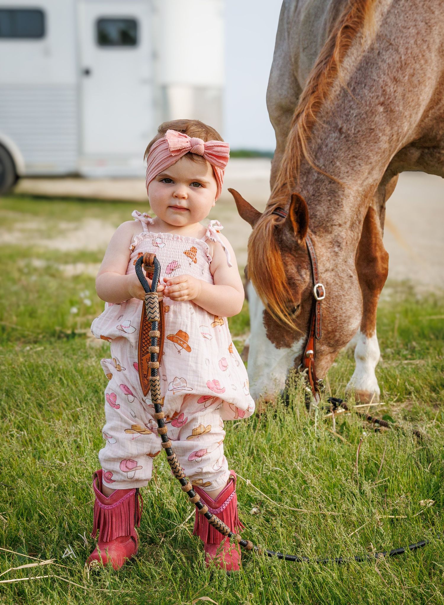 Toddler girl in pink outfit and cowboy boots holds horse lead rope beside grazing brown horse outdoors.
