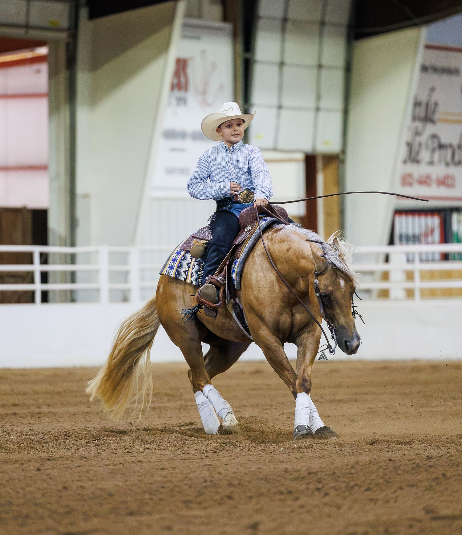Young rider in white cowboy hat competes on a palomino horse in an indoor Western equestrian arena.