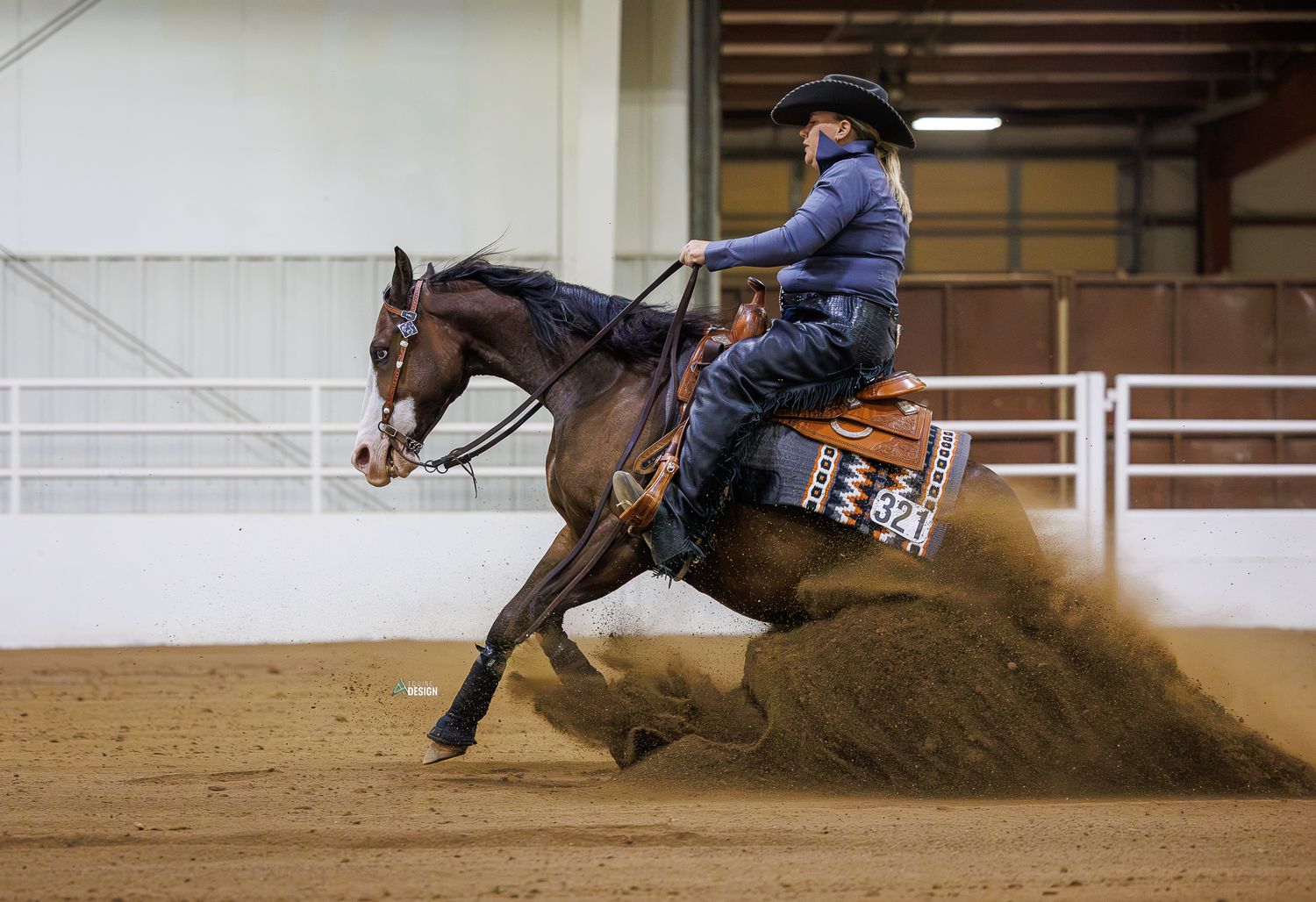Rider in blue performs a sliding stop on a brown horse in an indoor reining arena, kicking up dirt.