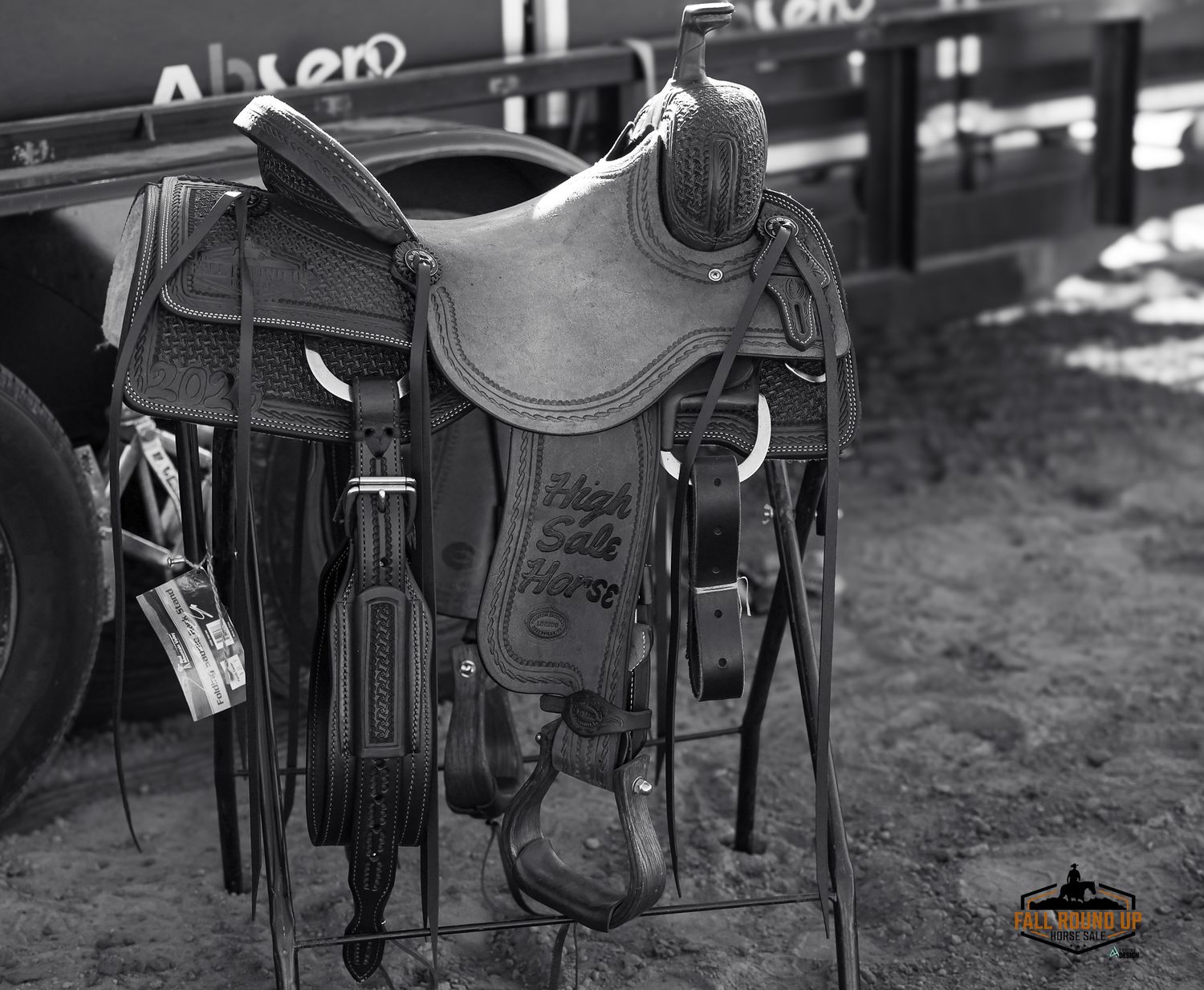 Black and white photo of a leather Western saddle resting on a metal stand at a rodeo event.