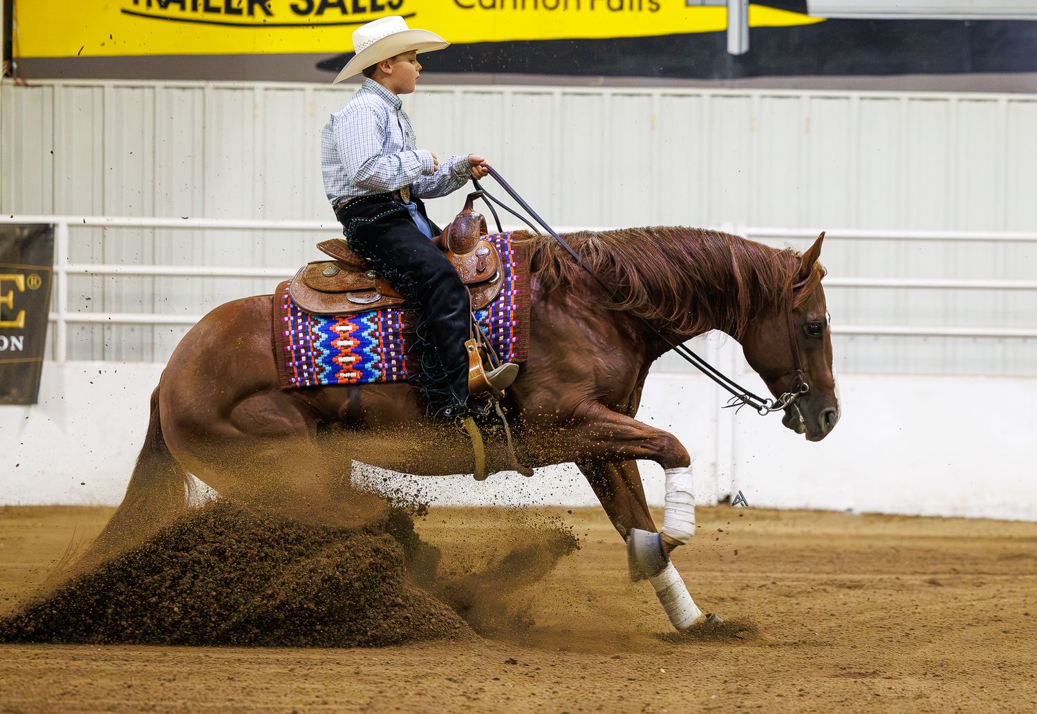 Cowboy riding a chestnut horse performing a sliding stop in an indoor reining competition arena.