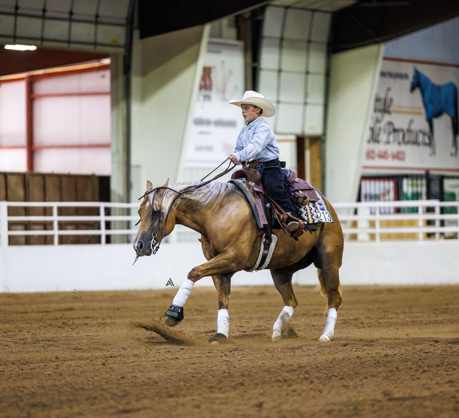 A young rider in a white cowboy hat competes on a palomino horse in an indoor Western equestrian arena.