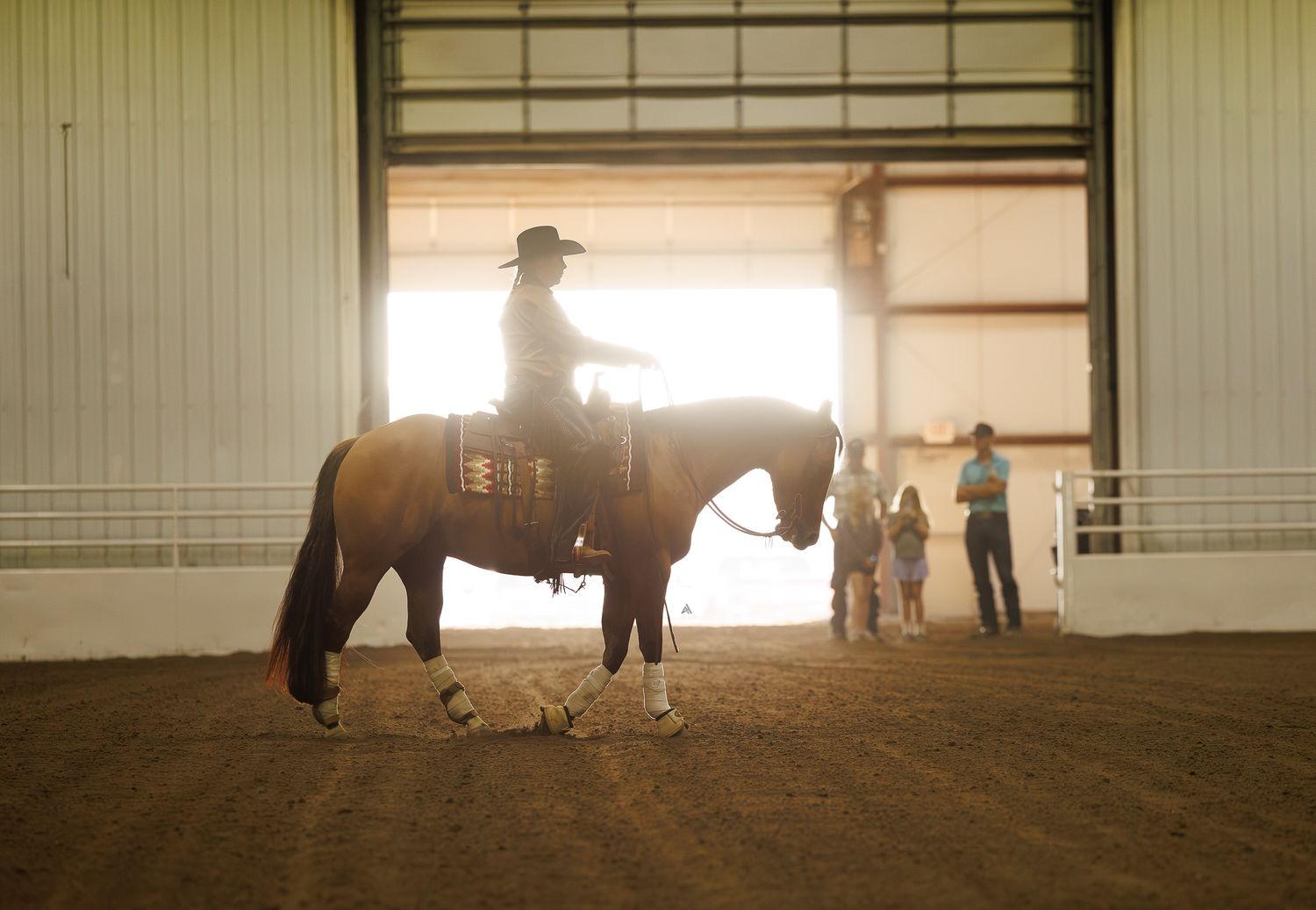 A cowboy rides a brown horse inside a sunlit indoor arena while children watch nearby.