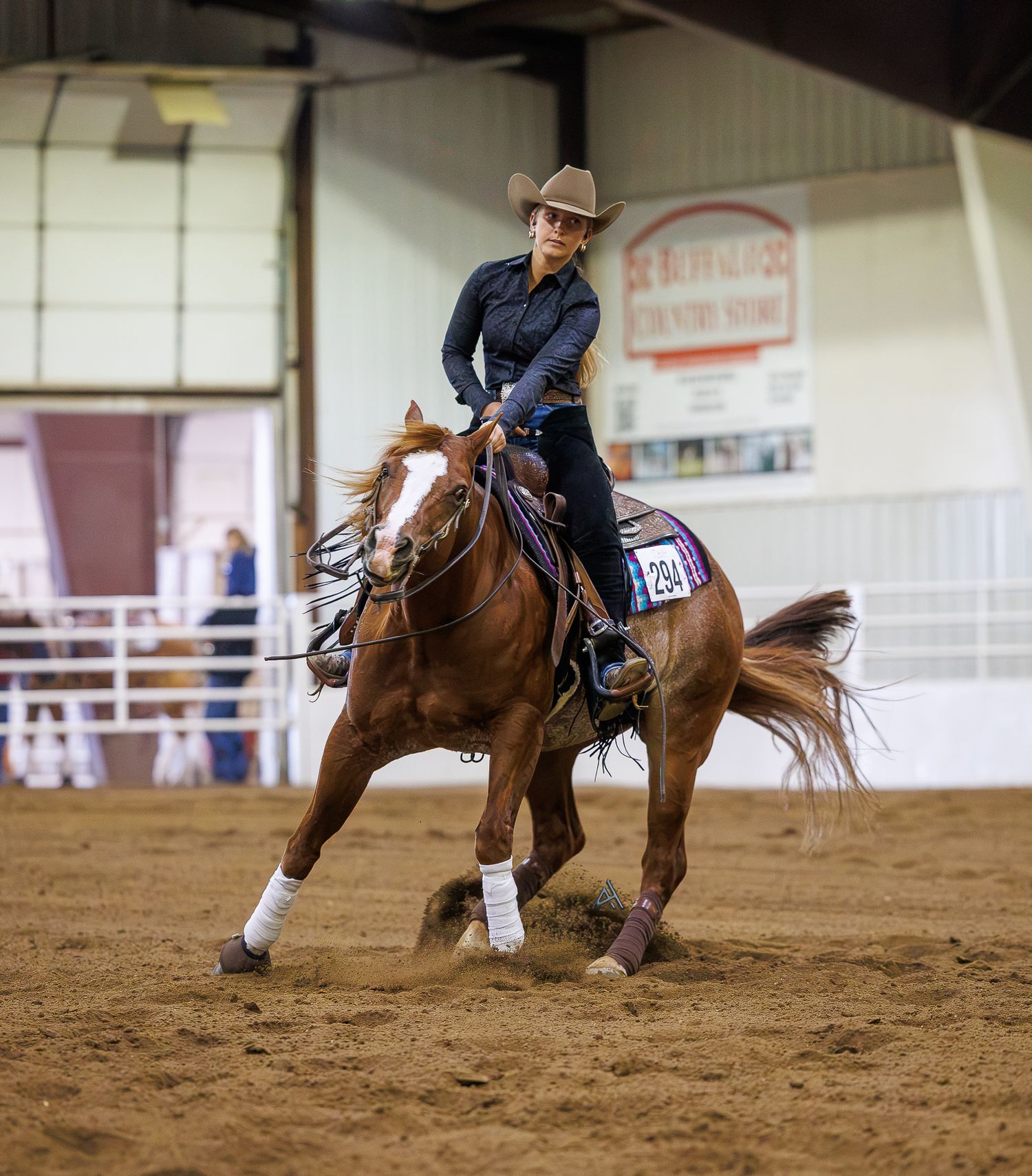 A female equestrian in a cowboy hat rides a chestnut horse during an indoor Western riding competition on a dirt arena.