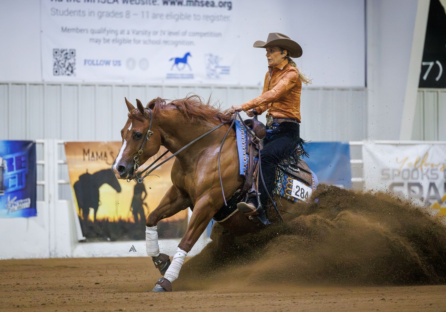 A rider in an orange shirt competes on a chestnut horse during a reining competition in an indoor arena.