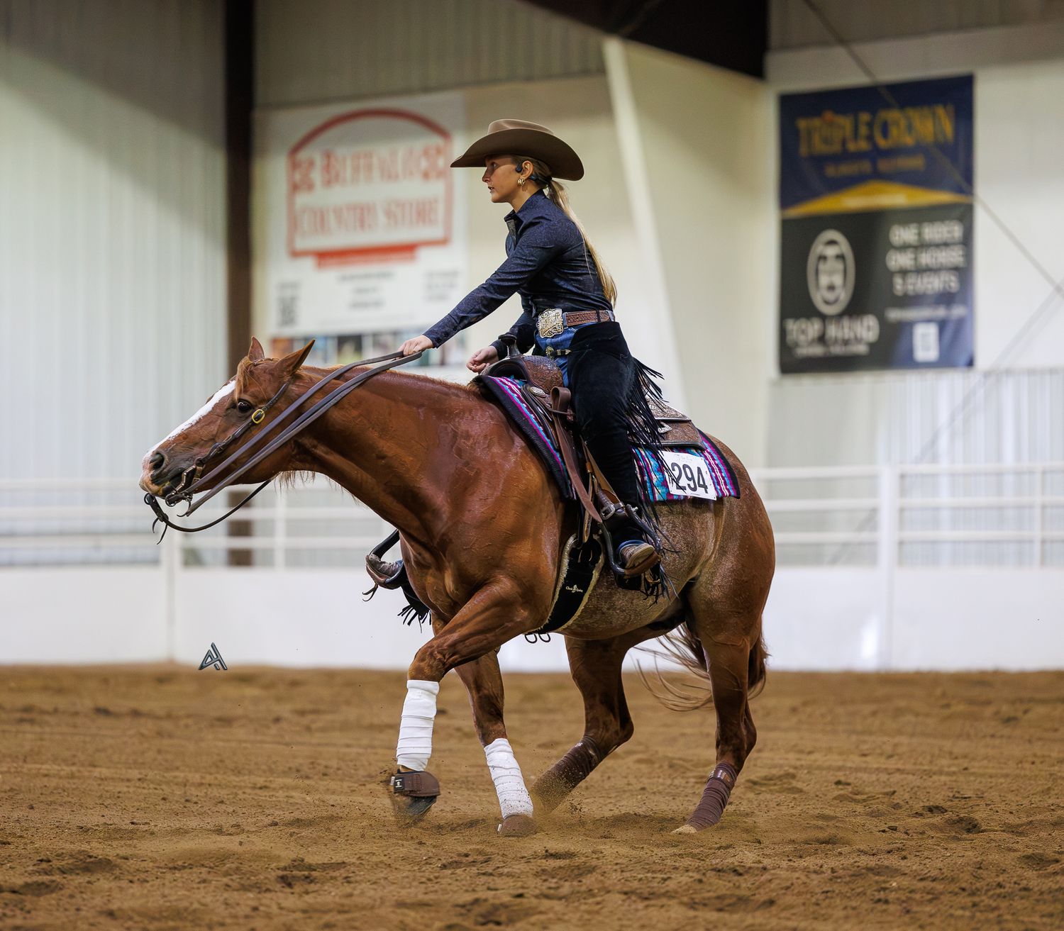 A female equestrian rider in a navy outfit competes on a chestnut horse in an indoor western reining arena.