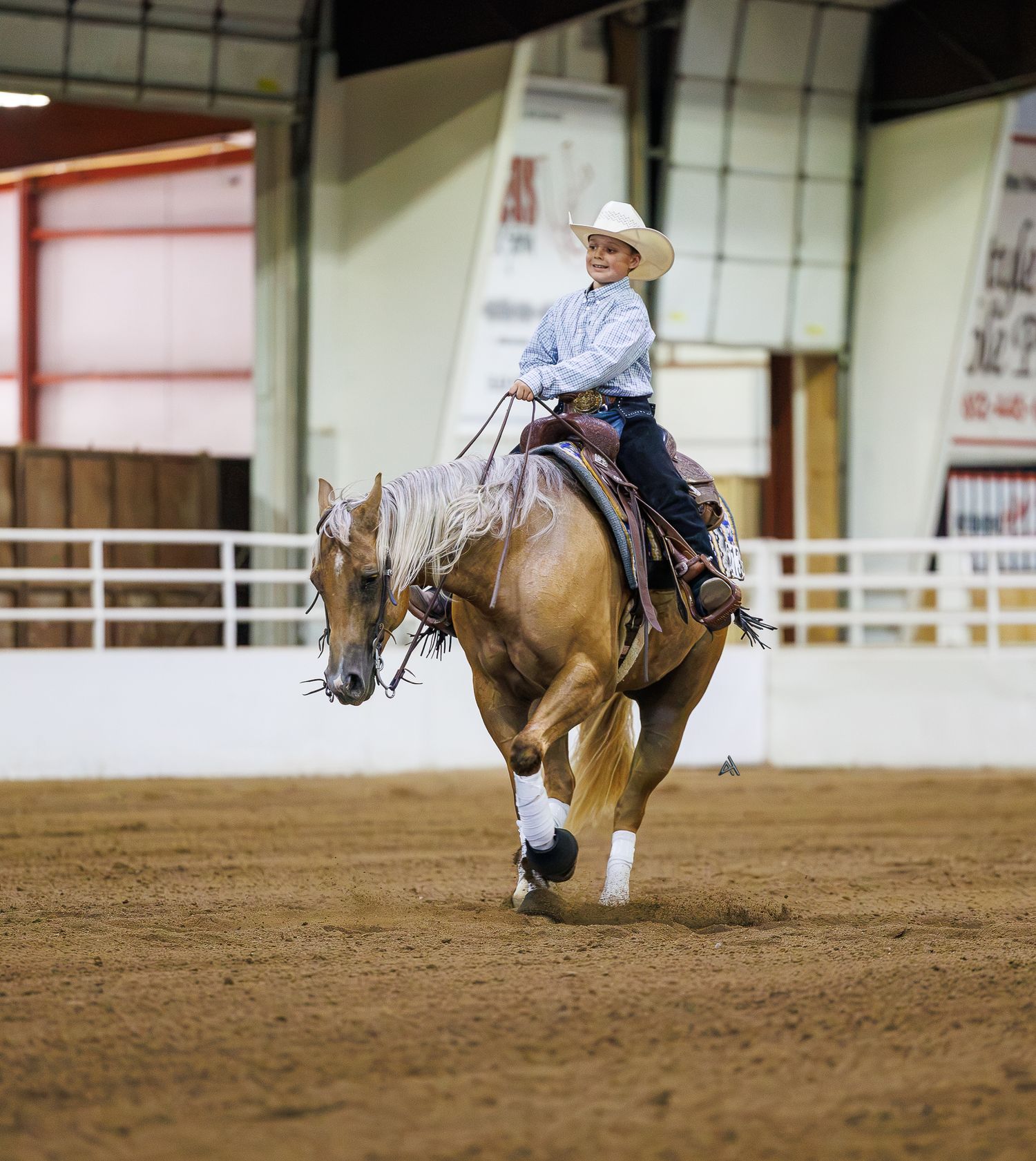 A cowgirl in a white hat rides a palomino horse in an indoor arena during a western equestrian competition.