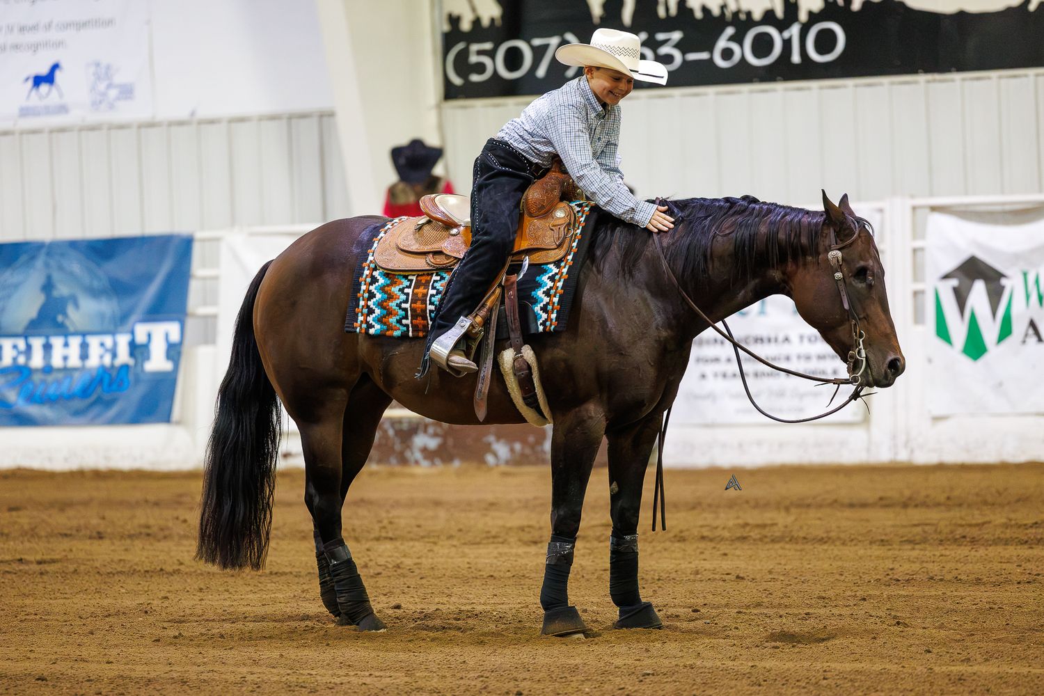 A young rider in western attire sits atop a dark horse in an indoor equestrian arena during a competition.