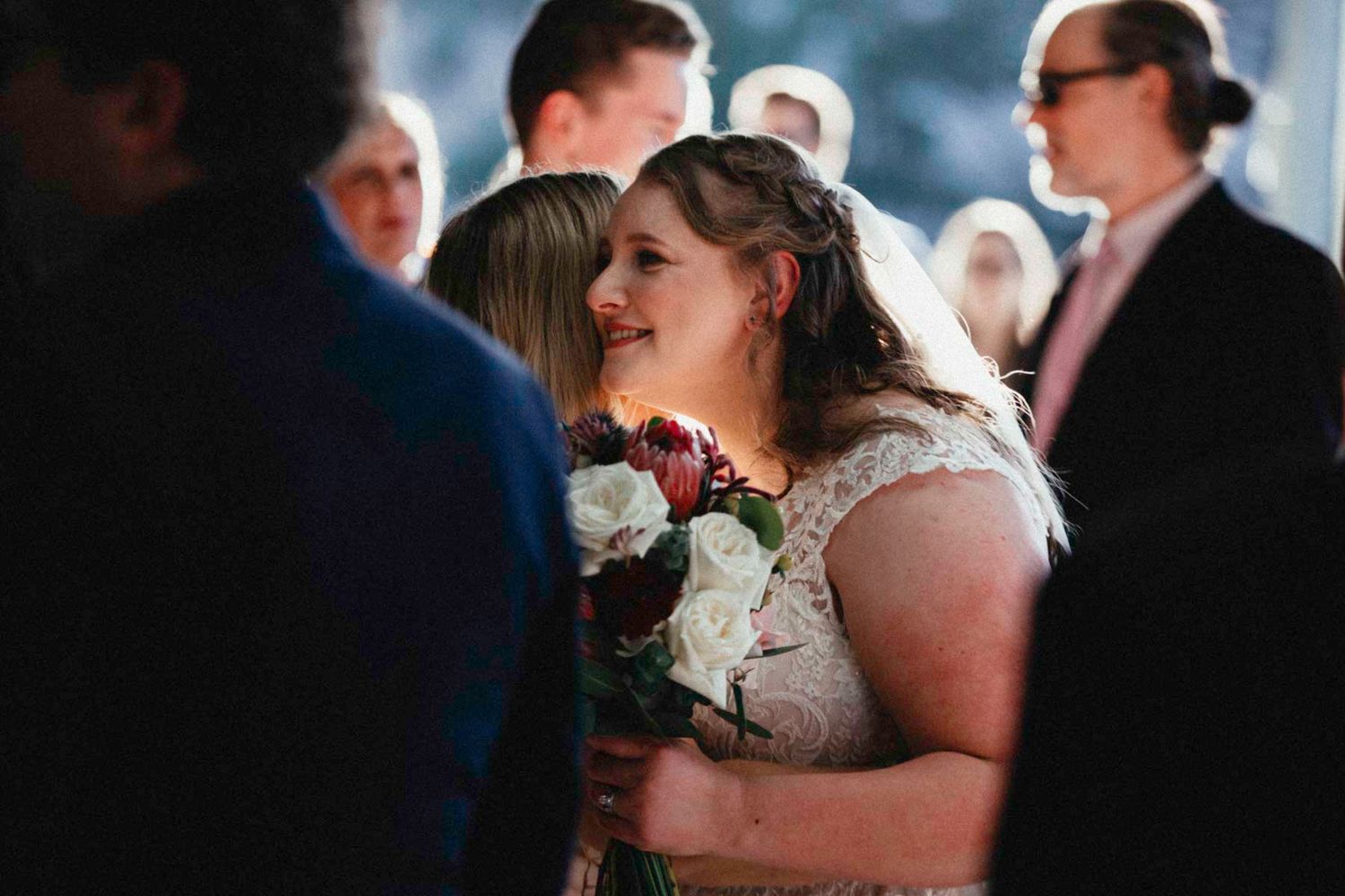 A bride in a white lace dress smiles while holding a bouquet during an indoor wedding ceremony.