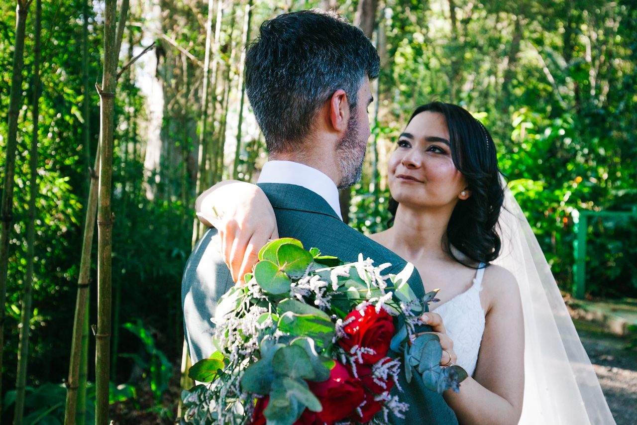A couple shares an intimate moment in a sunlit forest setting with a red and white wedding bouquet.