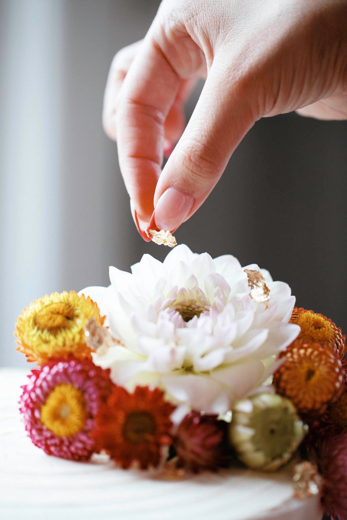 Hands delicately arranging colorful dried straw flowers and white chrysanthemums in a row on a white surface.