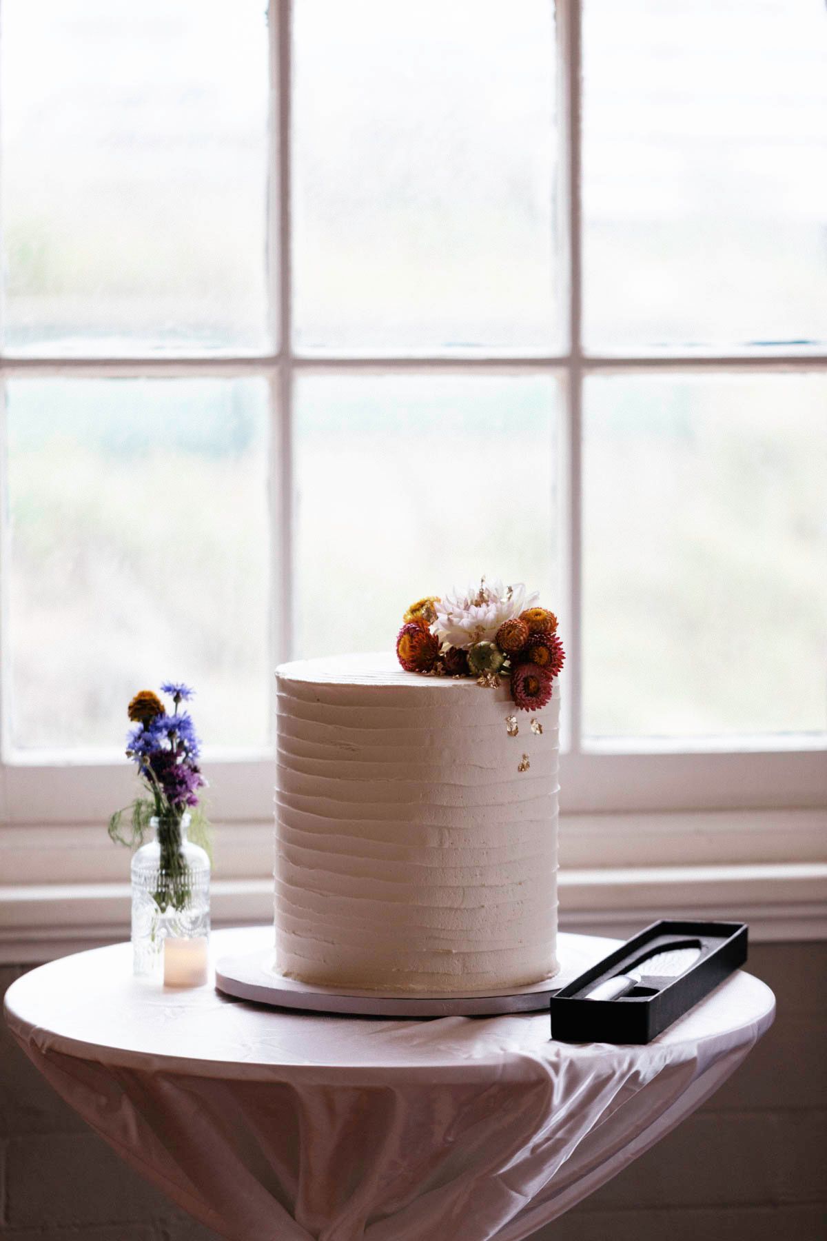 White wedding cake with horizontal texture lines sits on display near a window with natural lighting.