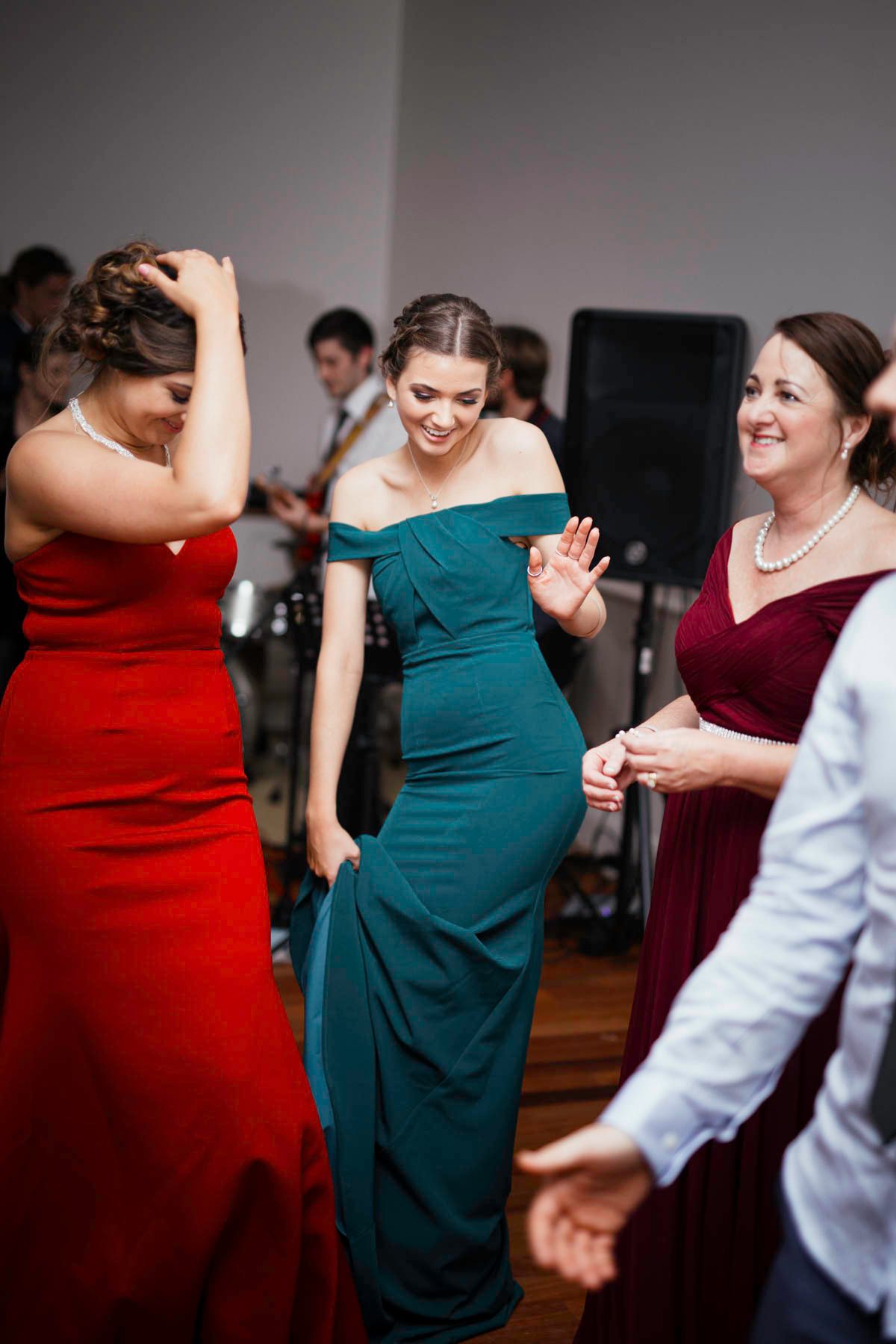 Wedding guests in red and teal dresses dance together joyfully at a reception celebration.