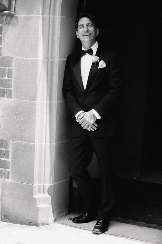 Person in formal black tuxedo standing in a stone doorway.