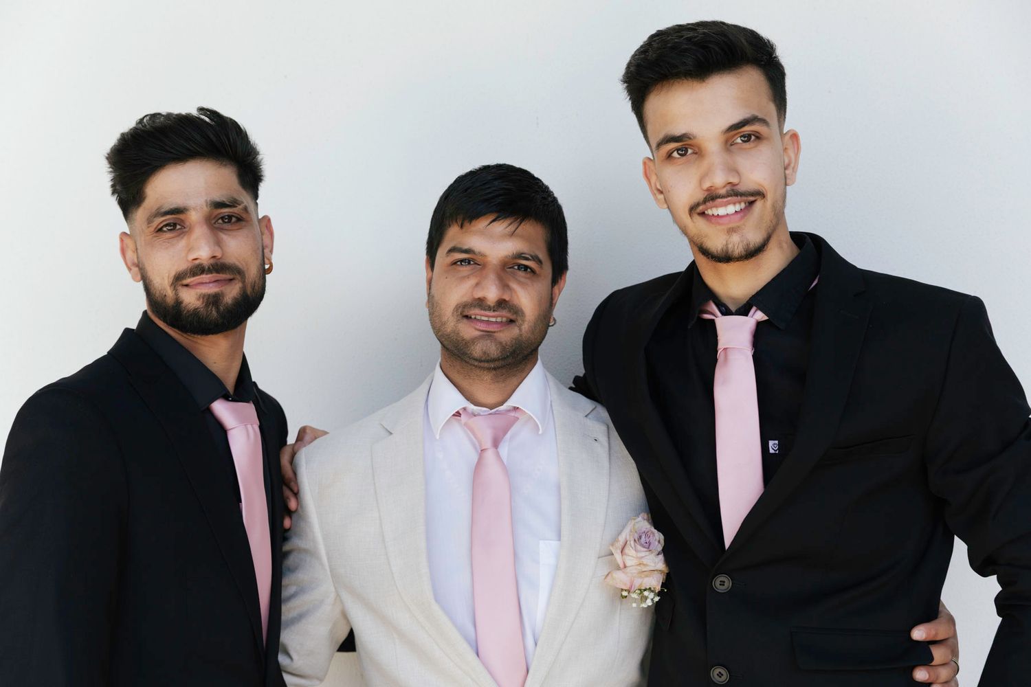 Three men in black suits with pink ties stand together smiling against a white background.