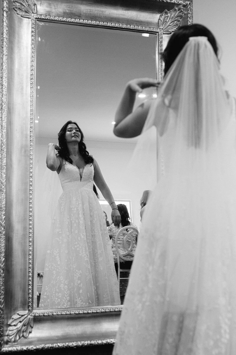 Black and white photo showing a bride trying on wedding dresses in a bridal boutique with ornate mirrors.