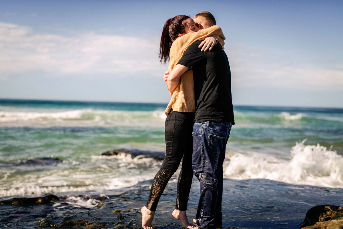 Romantic couple embraces on a rocky beach with crashing waves and ocean in the background.