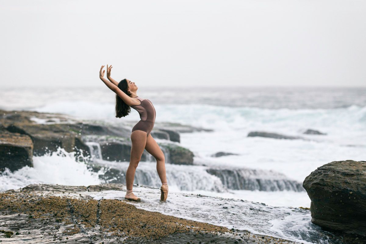 Sabela in ballet leotard posing gracefully on rocky coastal cliffs as ocean waves crash against the shore.
