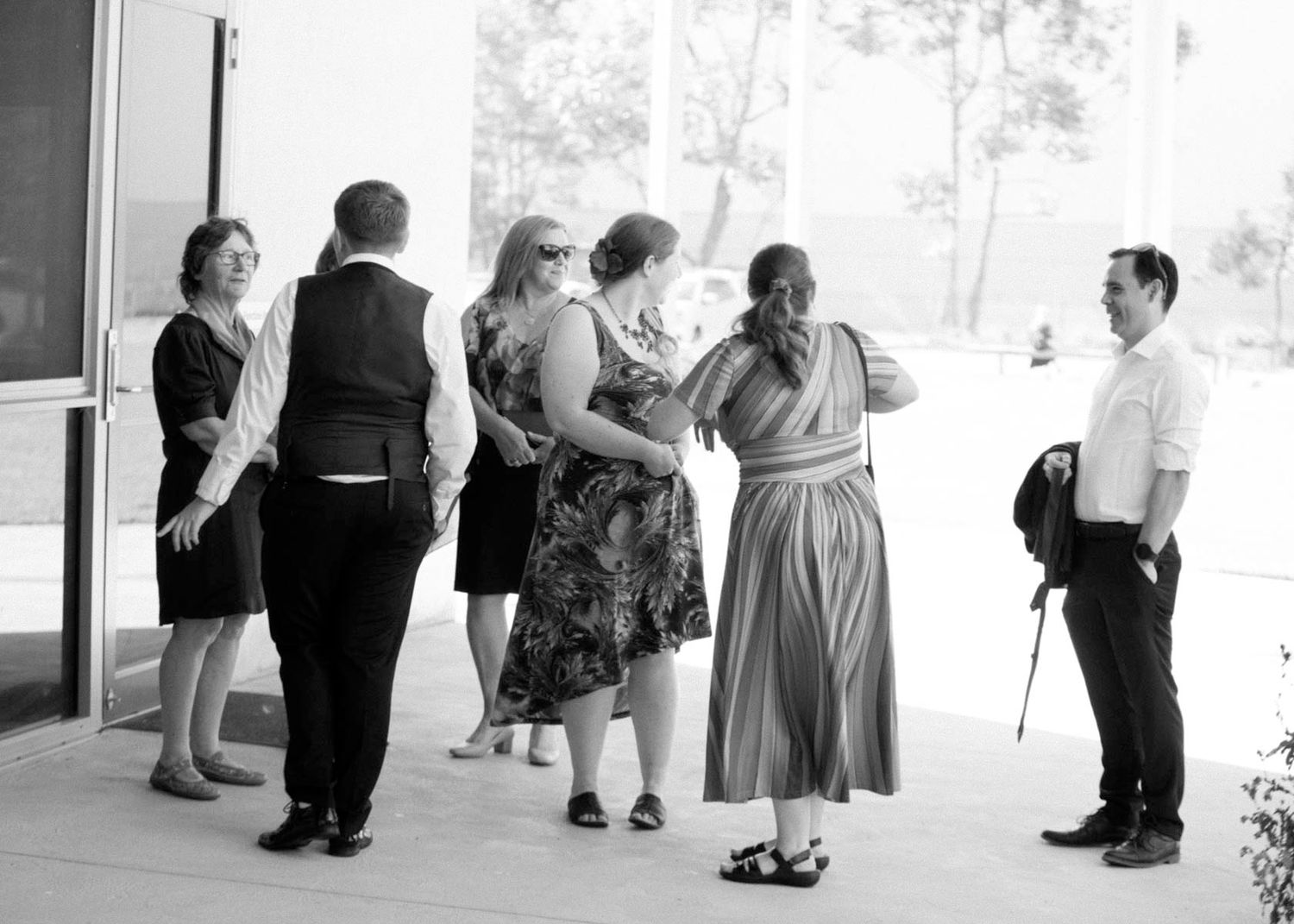 A black and white photograph shows a group of people gathered and conversing on a covered porch or walkway.