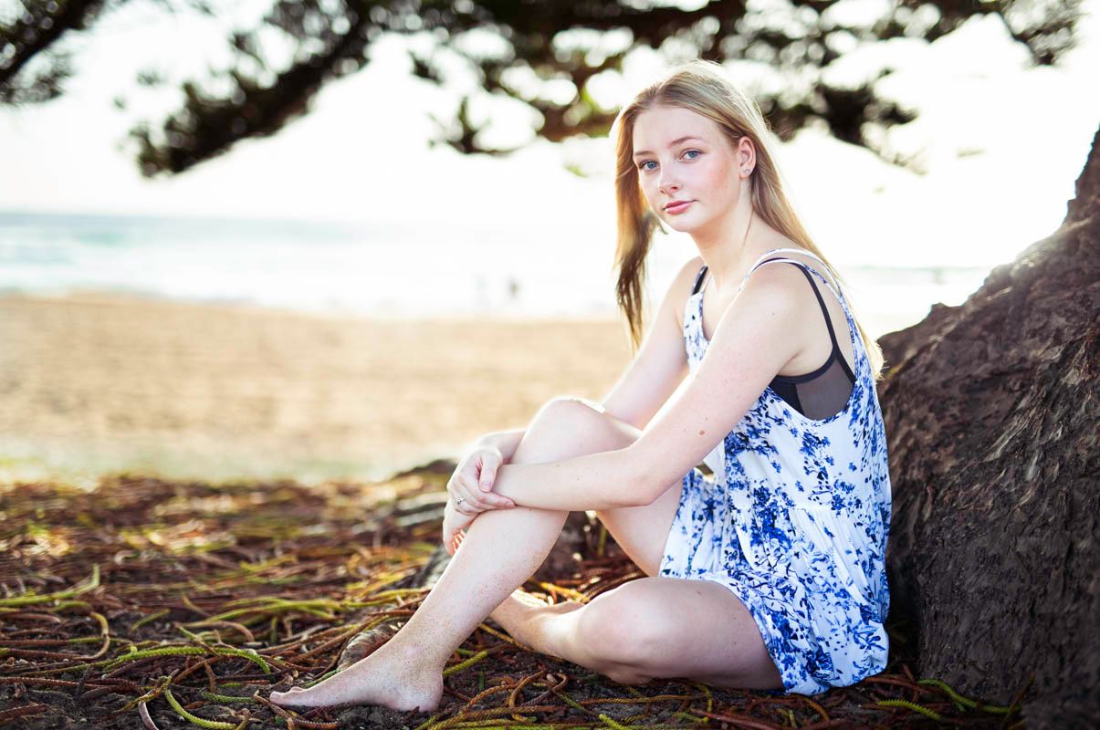 A blonde girl in a blue floral dress poses on a beach during golden hour creating artistic outdoor portraits.