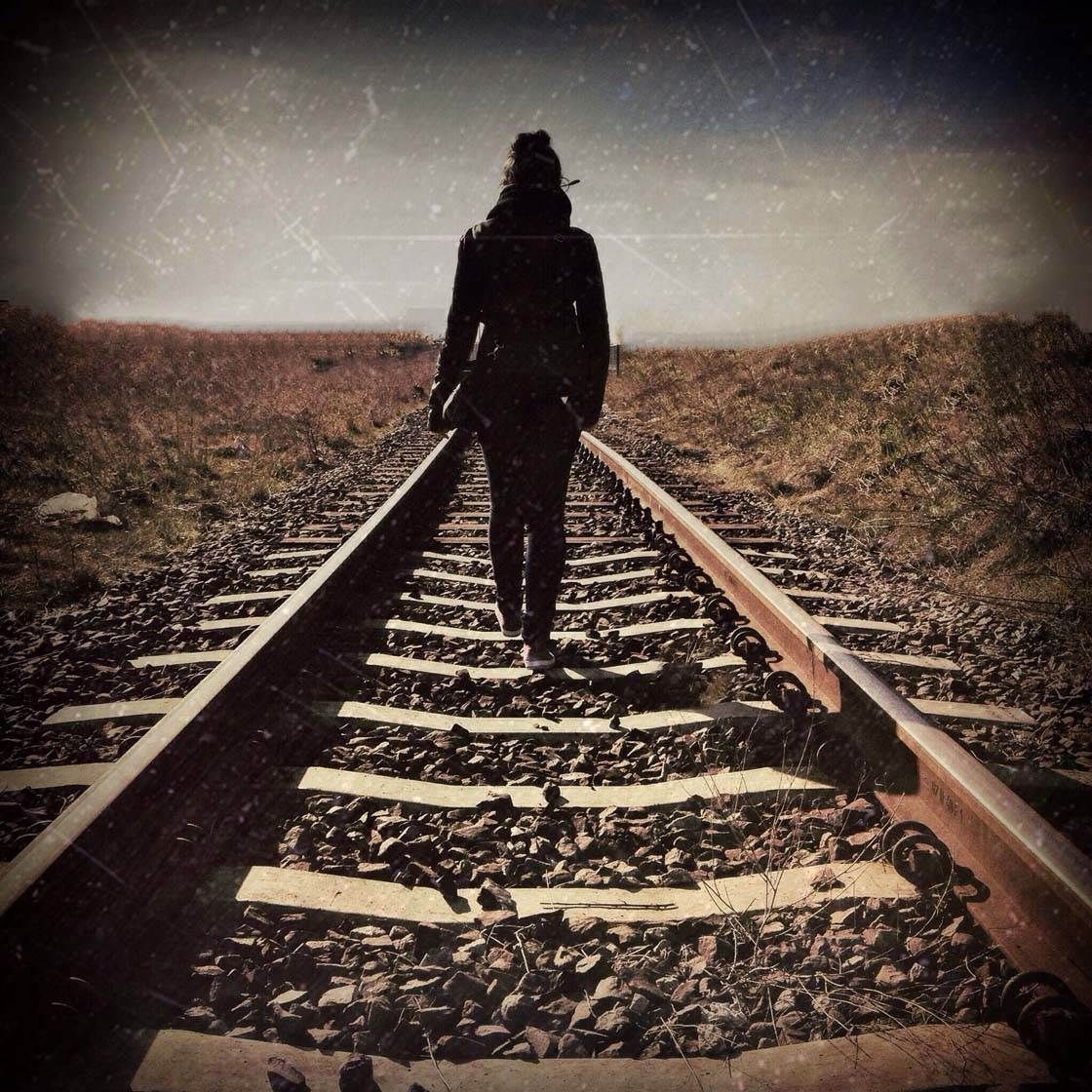 A lone person walks along empty railroad tracks into the distance, surrounded by a vast, desolate landscape.