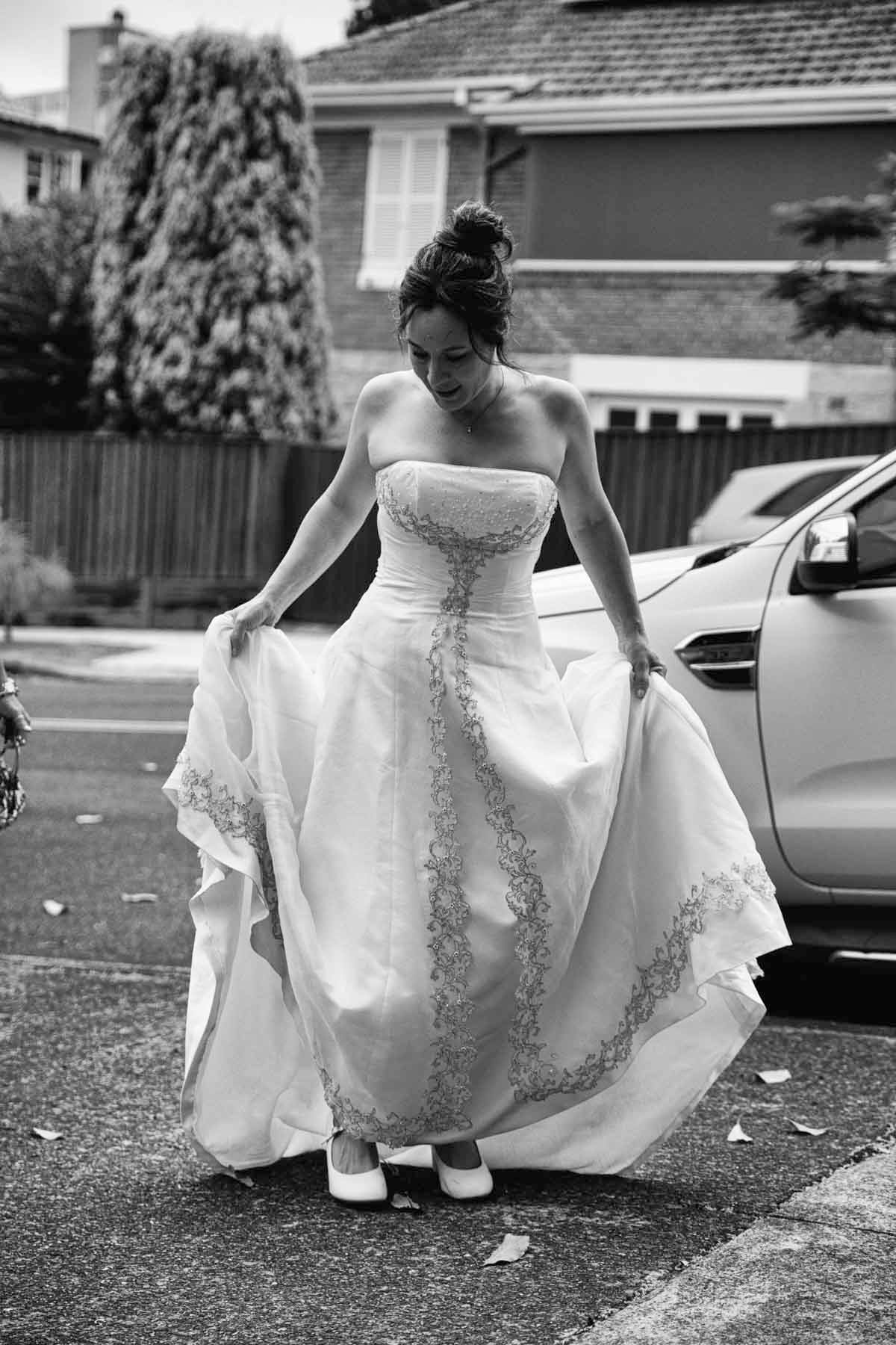 A bride in a strapless white wedding gown with embroidered details lifts her dress while standing in a driveway.
