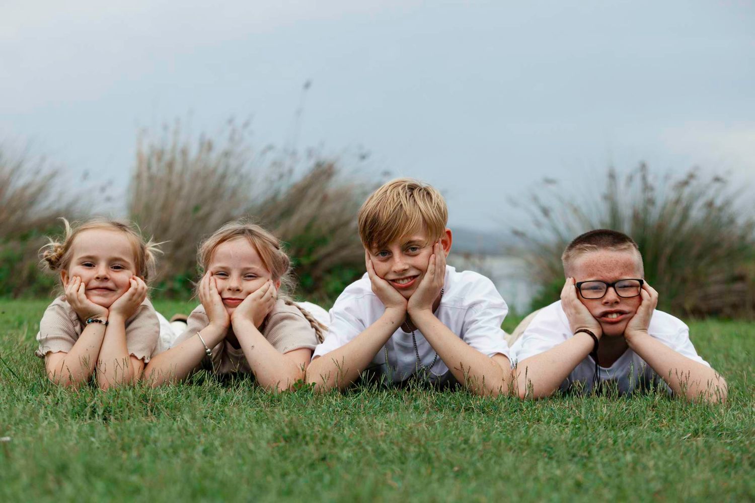 Four children lying on green grass in a row with their heads propped up on their hands.