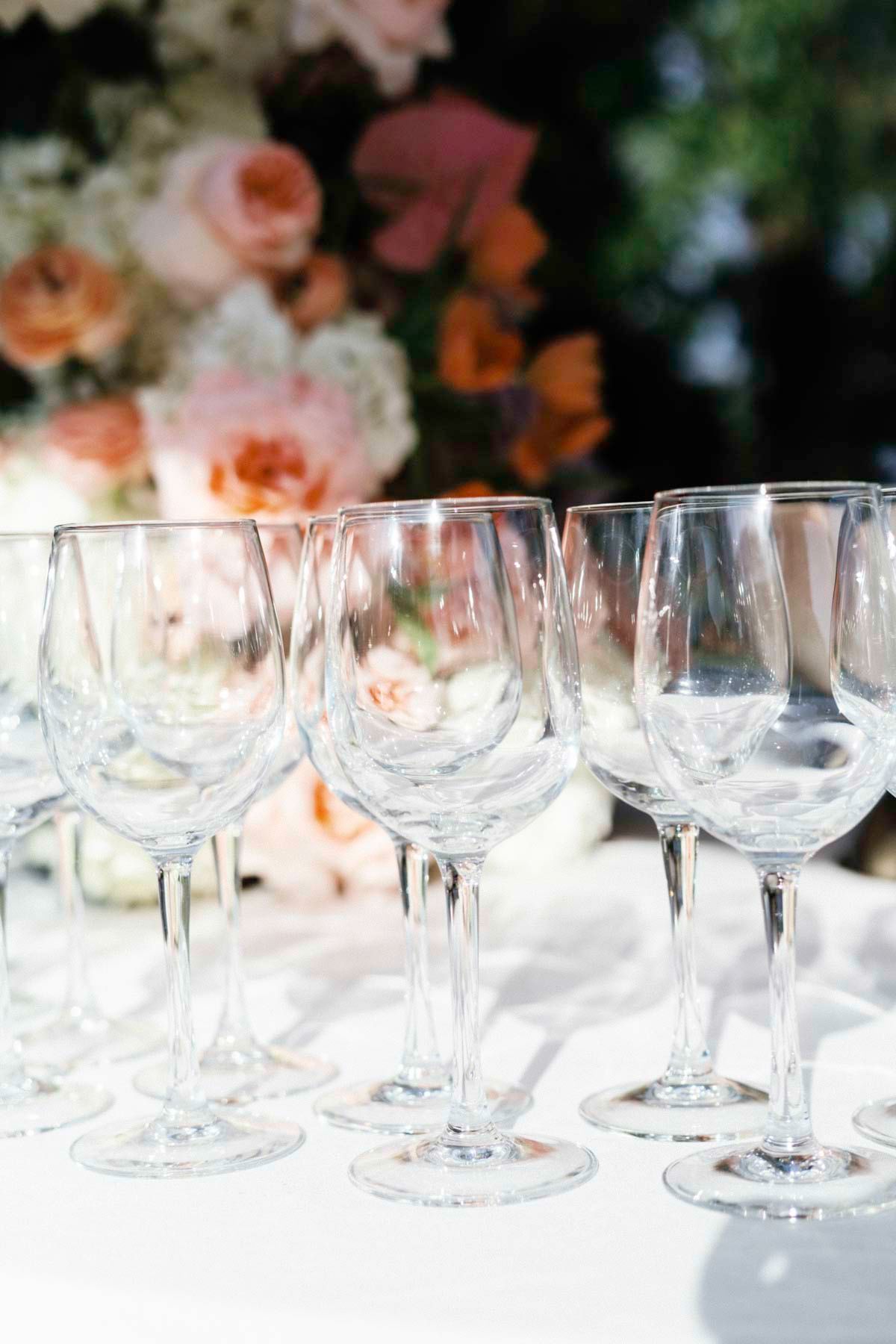 Empty wine glasses lined up on white tablecloth with soft pink roses in background creating elegant table setting.