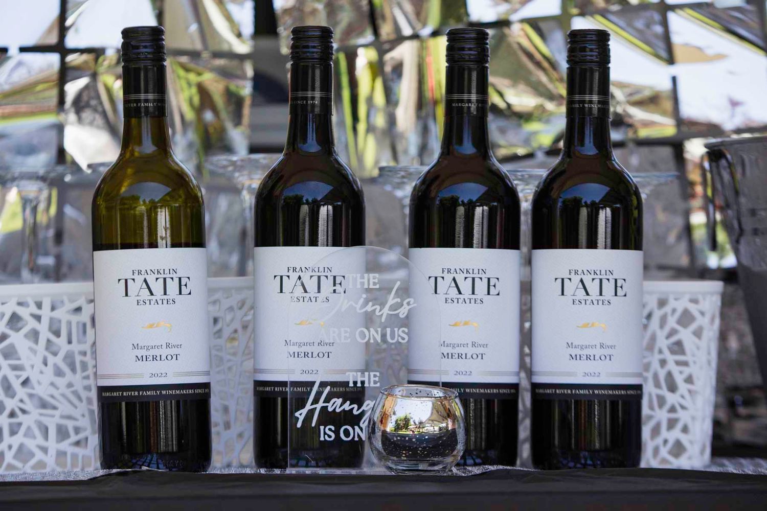 Rows of Tate brand wine bottles displayed on wooden tables at an outdoor event with palm trees in background.