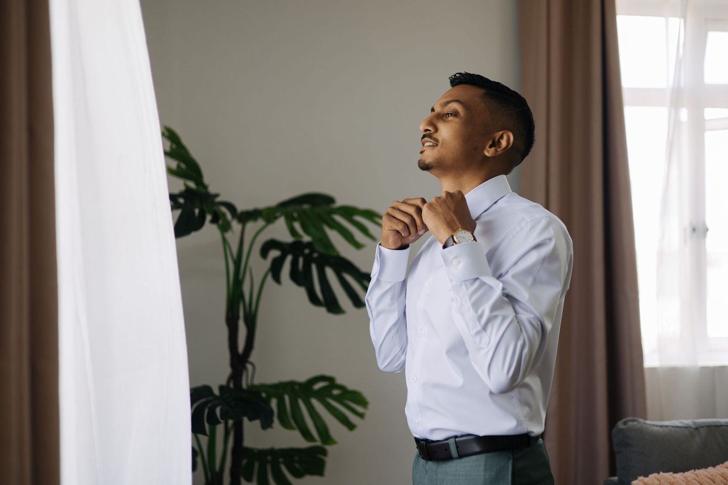 Stylish groom in white dress shirt buttoning his collar while standing near a window with tropical plant in background.