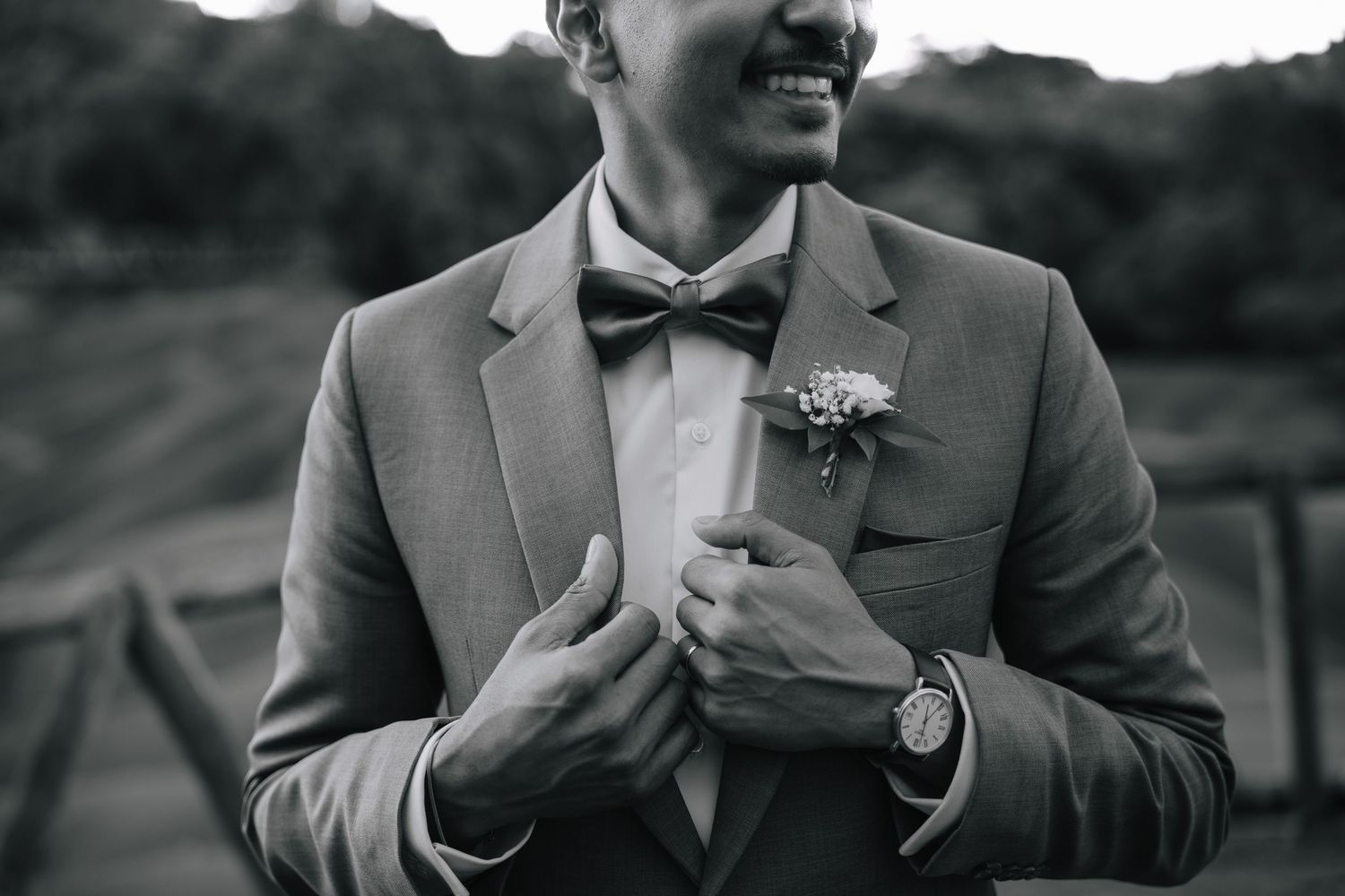Black and white photo of smiling groom in gray suit with bow tie and boutonniere adjusting jacket.