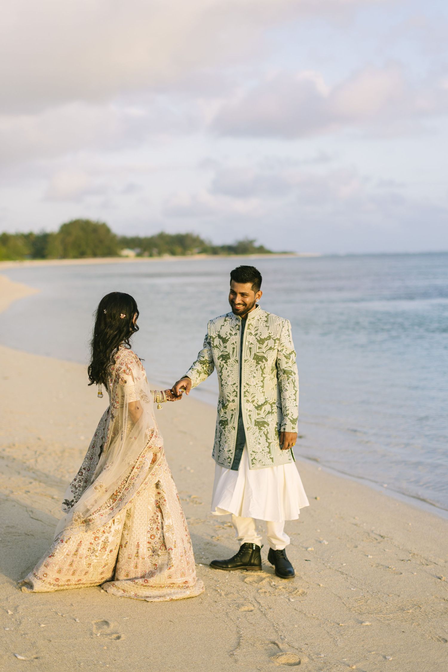 An Indian couple in traditional wedding attire hold hands and face each other on a beautiful sandy beach.