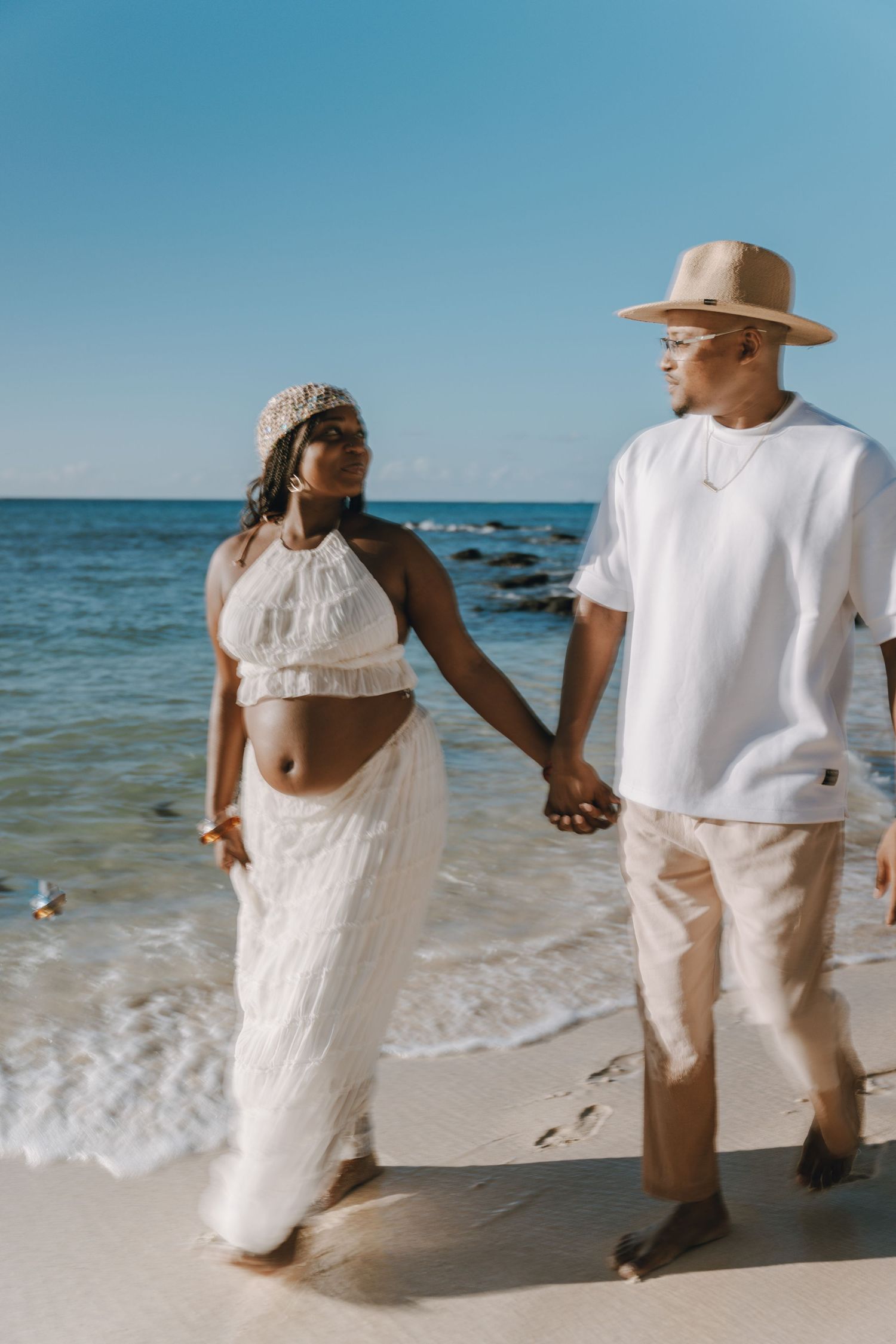 Pregnant couple walking hand in hand on a sunny beach, woman in white two-piece outfit, man in white shirt and straw hat.