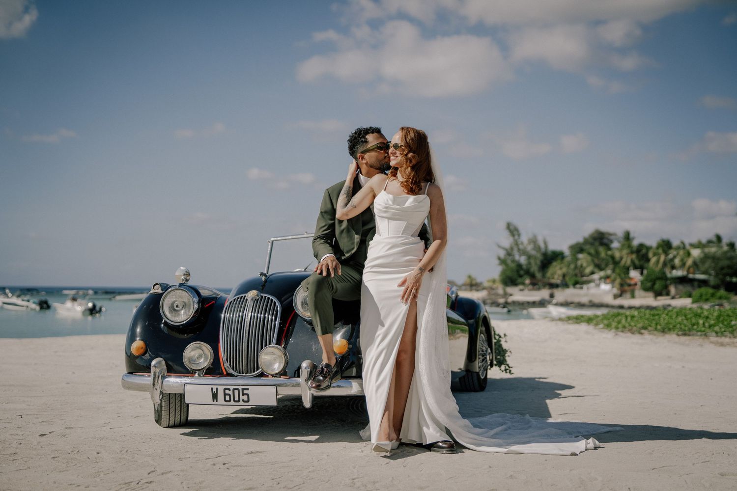 Bride and groom posing beside vintage blue car on tropical beach with clear blue sky backdrop.