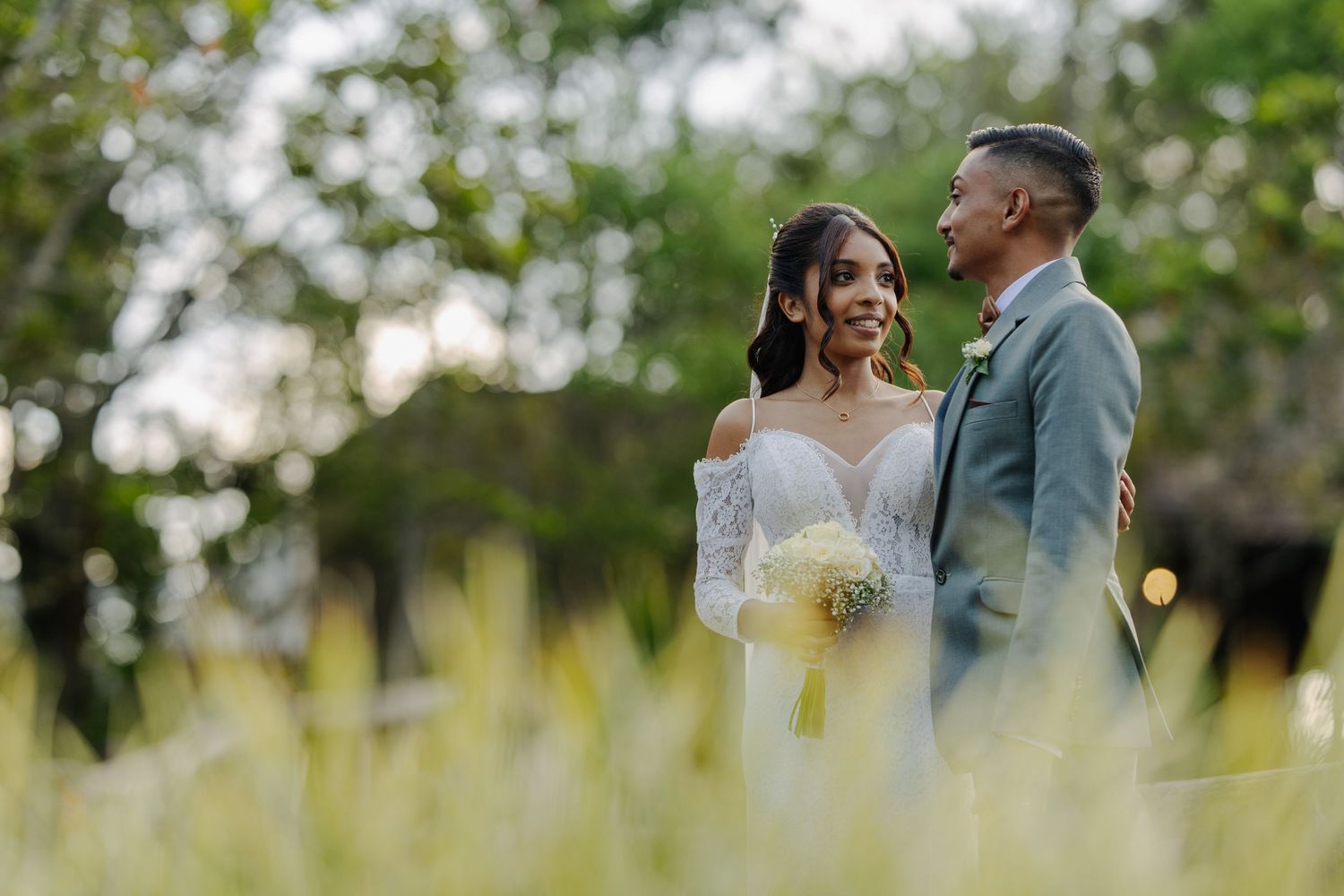 Bride and groom sharing a tender look outdoors at golden hour, bride holding yellow bouquet in lace wedding gown.